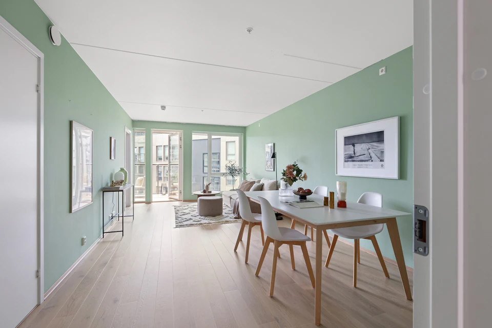 The image depicts a modern, open-concept living room and dining area with light green walls that reflect off the white ceiling. The room features a large window on the left wall, allowing natural light to flood the space. A wooden floor adds warmth to the contemporary setting. In the center of the room is a white table surrounded by four chairs, suggesting an inviting atmosphere for socializing or dining. On one side of the room, there's a couch and a coffee table with a vase filled with flowers on top. The walls are adorned with two framed pictures, adding character to the space.