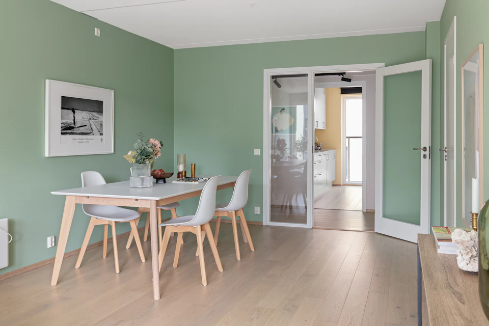 The image depicts a modern, open-concept living room with a large window that allows natural light to flood the space. The room features a white table surrounded by four chairs, two on each side and one at the head of the table. On the table are three vases filled with flowers, adding a touch of color and life to the room. A painting is hung above the table, contributing to the overall aesthetic. In the background, there's a kitchen area visible through an open door, suggesting that this space serves multiple functions within the home. The flooring consists of hardwood planks, which complement the white walls and create a cohesive design throughout the living room.