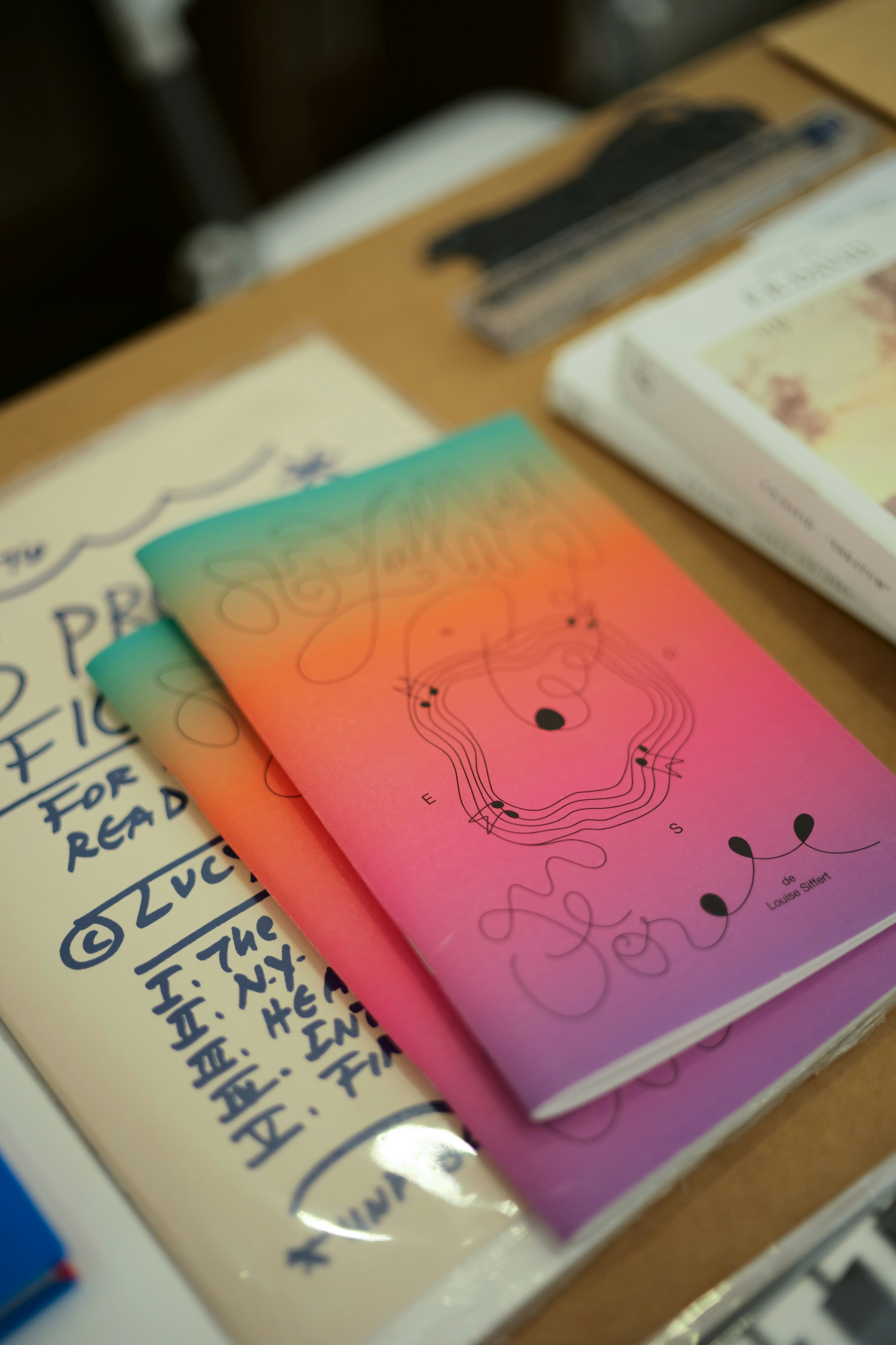 An open zine with a gradient cover lying on a desk among notebooks