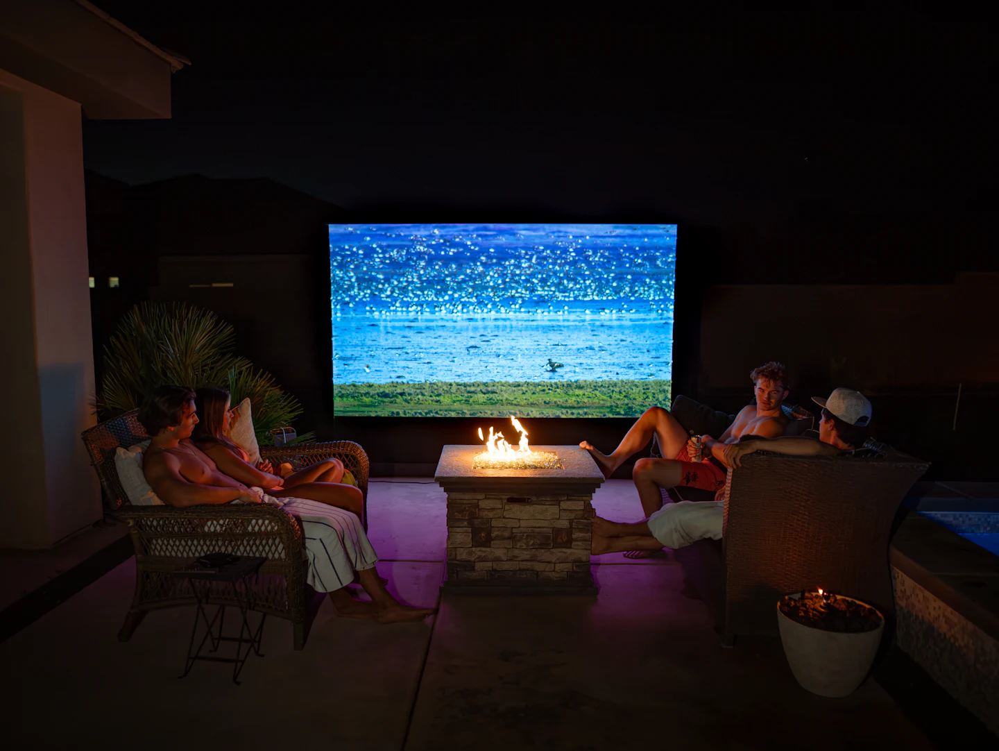 Friends relaxing around a fire pit while a large outdoor TV plays at night