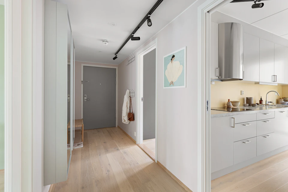 The image depicts a modern, open-concept living space with white walls and light wood flooring. The room features a gray door leading to a hallway on one side, while another hallway leads directly into the kitchen area. A large window allows natural light to flood in, illuminating the space. On the right wall, there is a painting of a woman's face hanging above the sink. In the center of the image, a wooden bench can be seen, and a coat rack stands nearby. The room also contains various pieces of furniture such as chairs and tables scattered throughout the area.