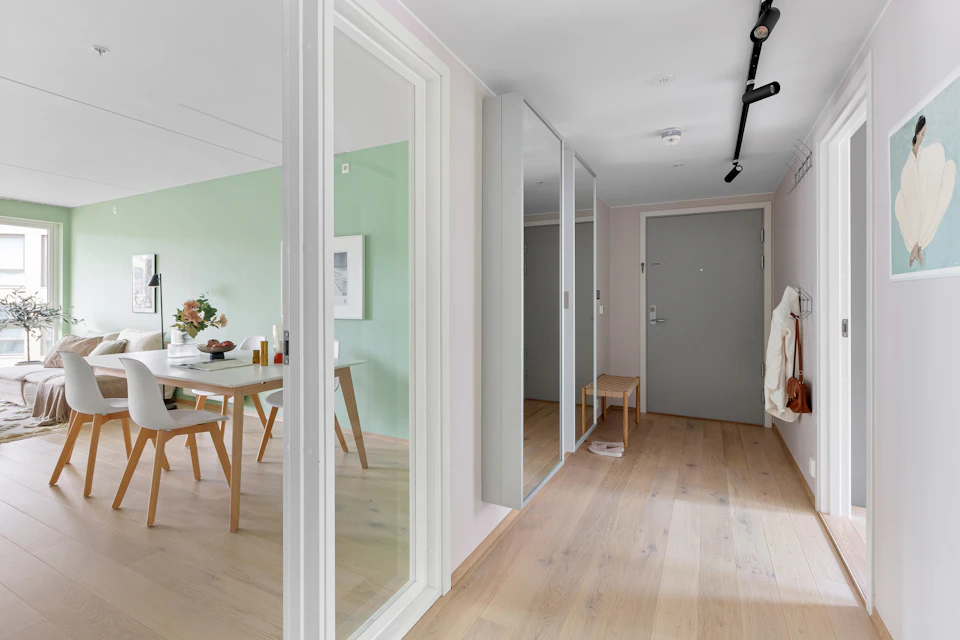 The image presents a modern and stylish living room with white walls, a gray carpeted floor, and wooden furniture. On one side of the room is a dining area featuring a table surrounded by four chairs, each chair painted in a different color - white, blue, green, and brown. The dining area is situated next to a hallway that leads to another room, which has a door with a coat rack on it.

In the background, there's a kitchen area with a sink and countertop, as well as a refrigerator. A vase of flowers sits on top of the table in the living room, adding a touch of nature and color to space.