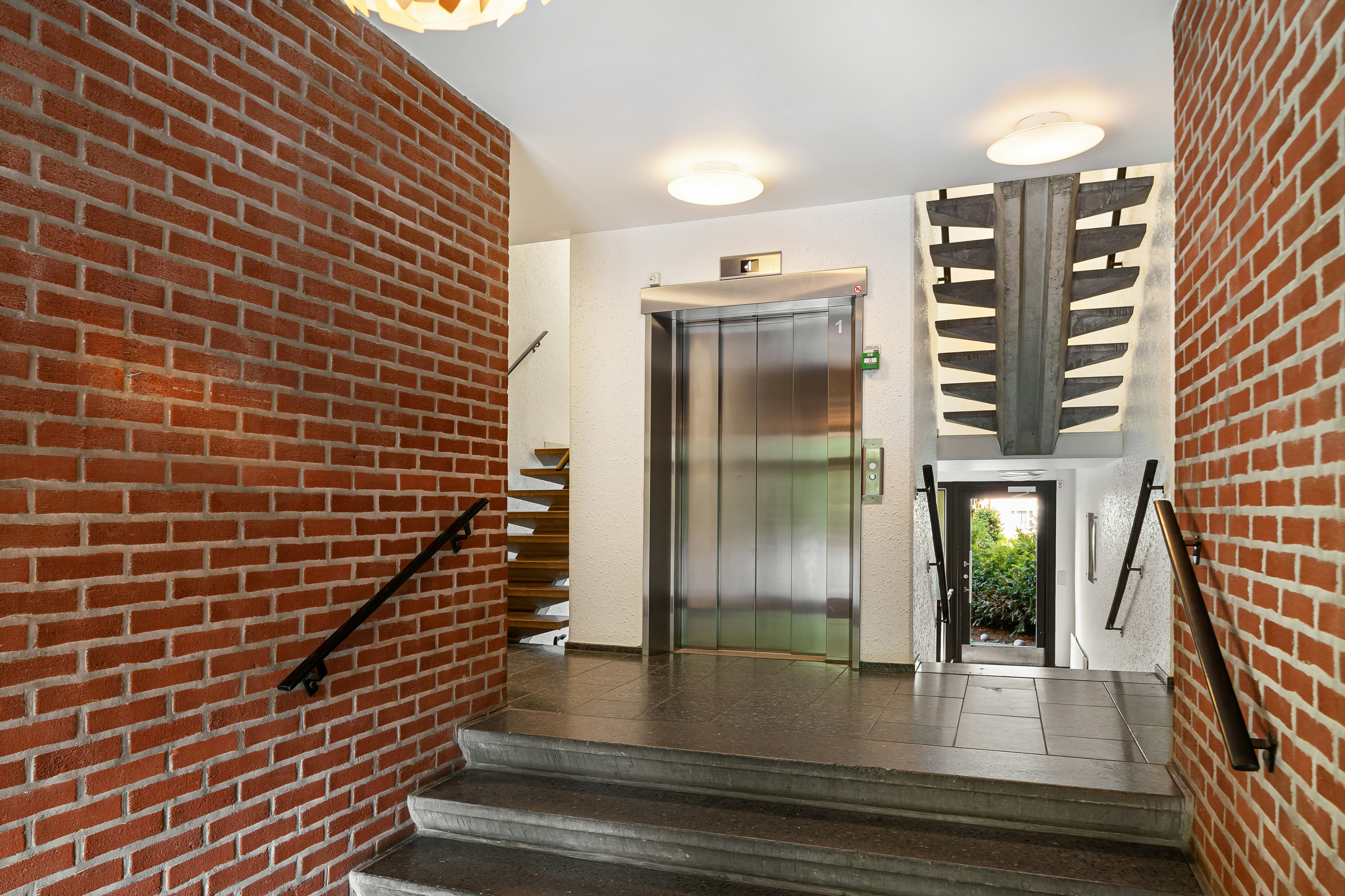 Modern building interior featuring a stainless steel elevator, red brick walls, and grey concrete stairs leading down to an outdoor view.