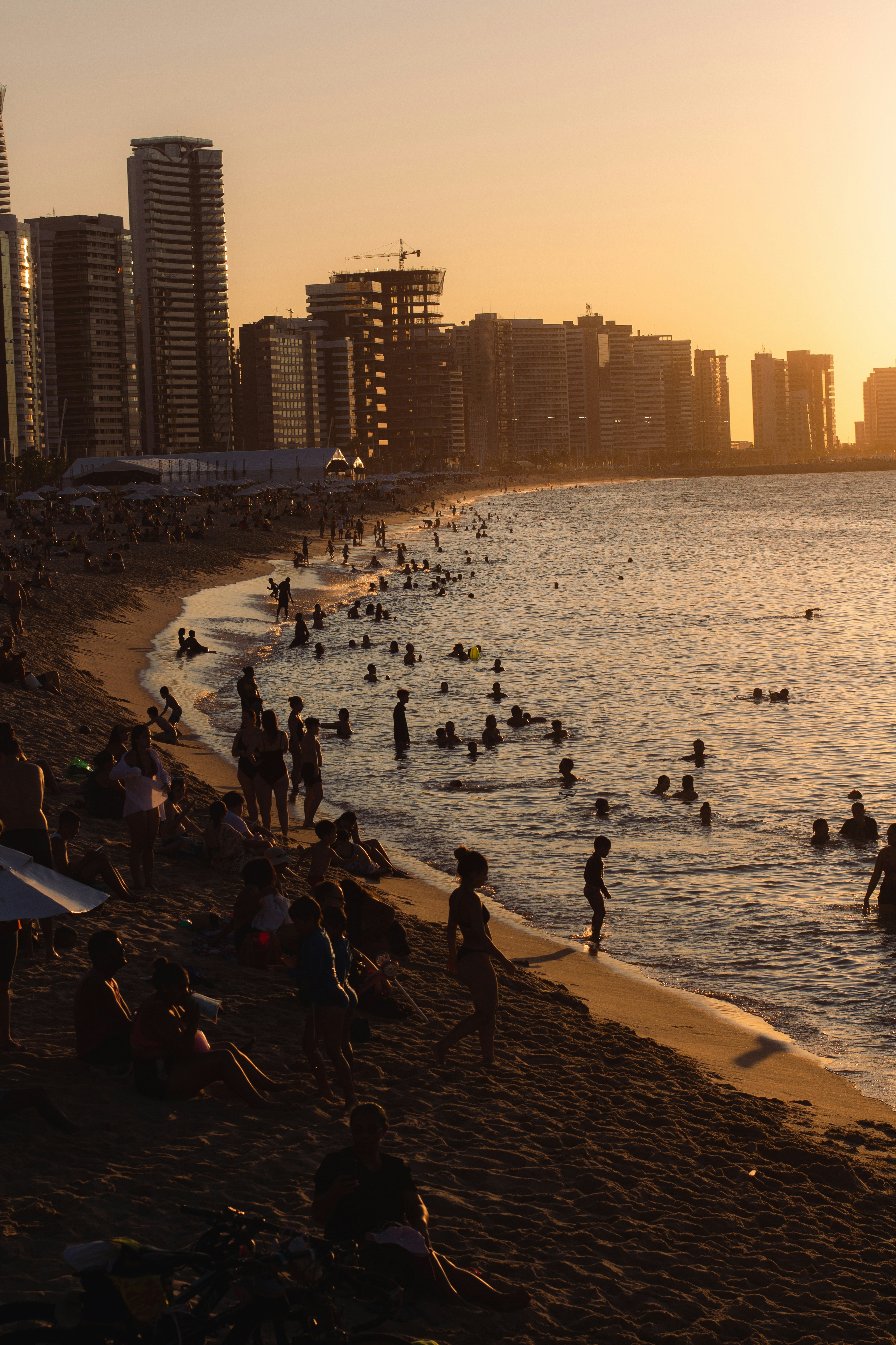 A crowded beach at sunset with people swimming in the water