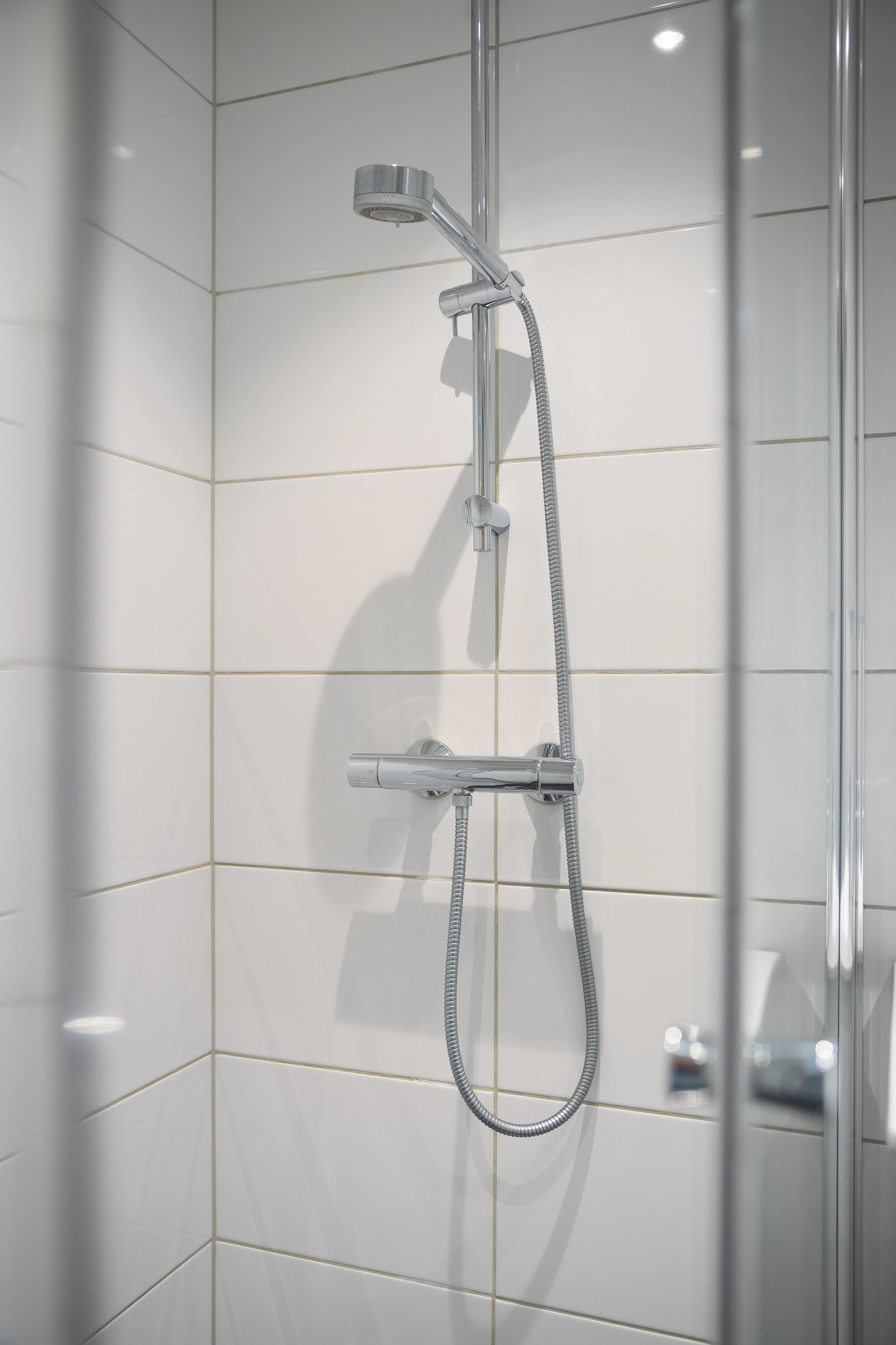 Close-up of a chrome handheld shower rail, mixer, hose and showerhead mounted on white tiled walls behind glass.