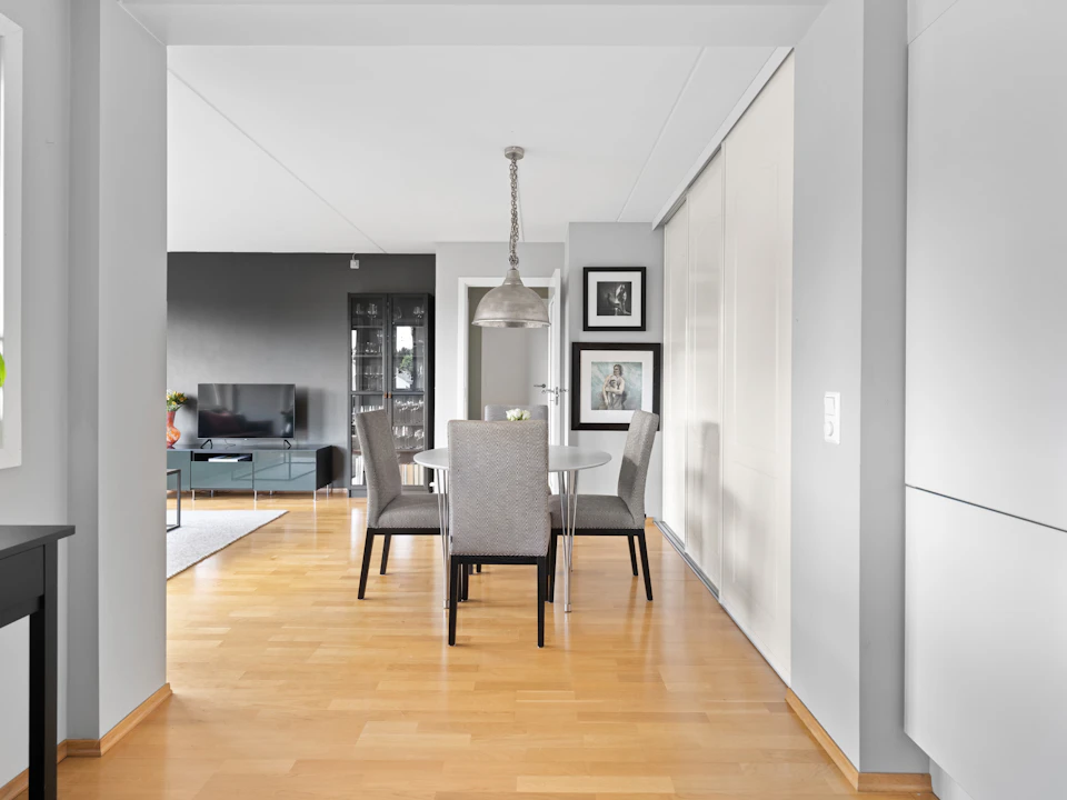 The image depicts a modern, open-concept living and dining room with white walls that reflect natural light from a large window on the left side. The floor is made of wood, adding warmth to the space. In the center of the room, there is a gray table surrounded by four chairs, suggesting an inviting atmosphere for gatherings or meals.

On one side of the room, there is a television and a bookshelf filled with various items, indicating that this area serves both entertainment and intellectual pursuits. The other side features white cabinets and a black door, providing ample storage space while maintaining the clean lines and simplicity of the design.