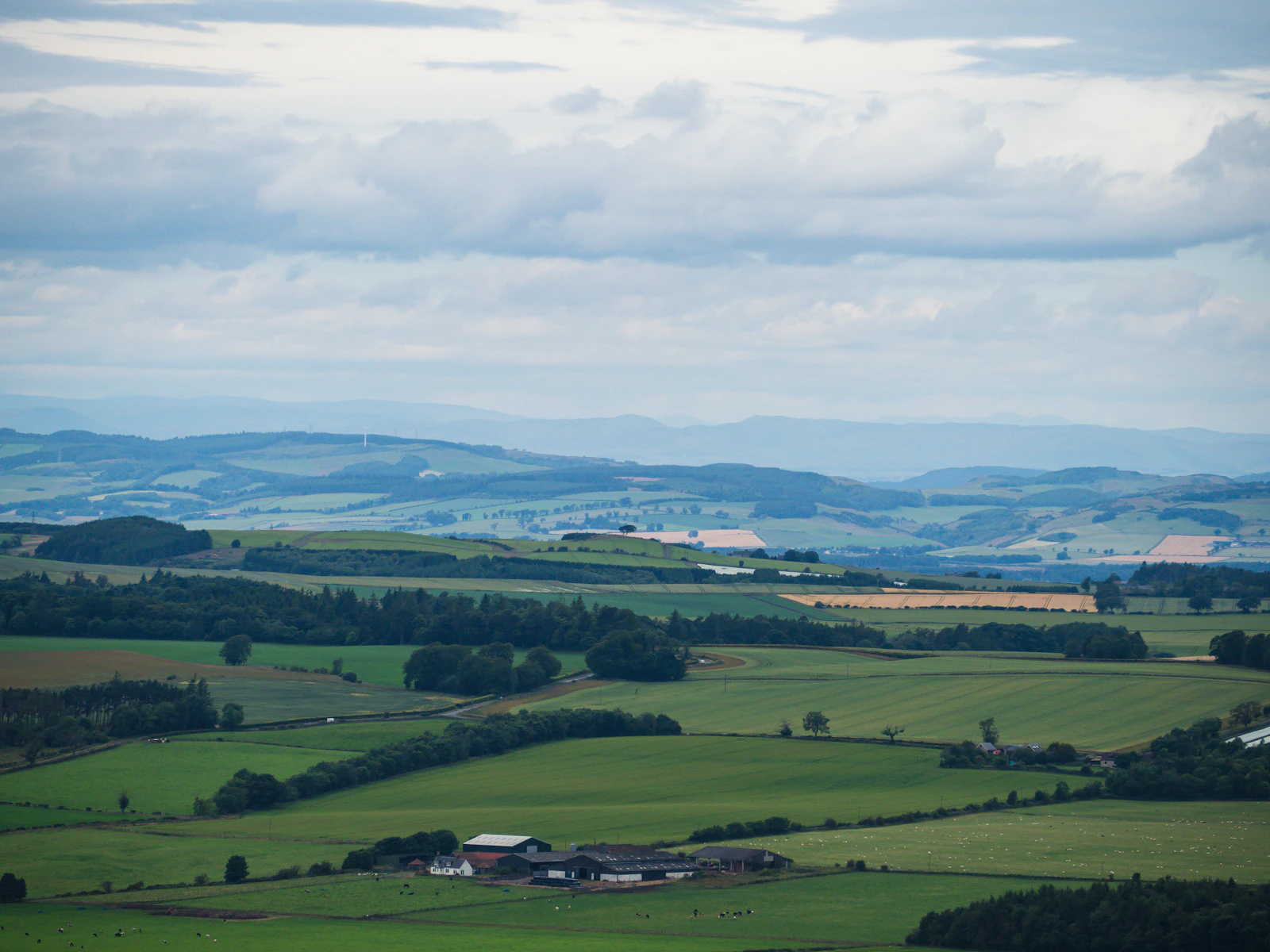 A broad agricultural valley seen from a hilltop, with fields, tree lines and distant ridges