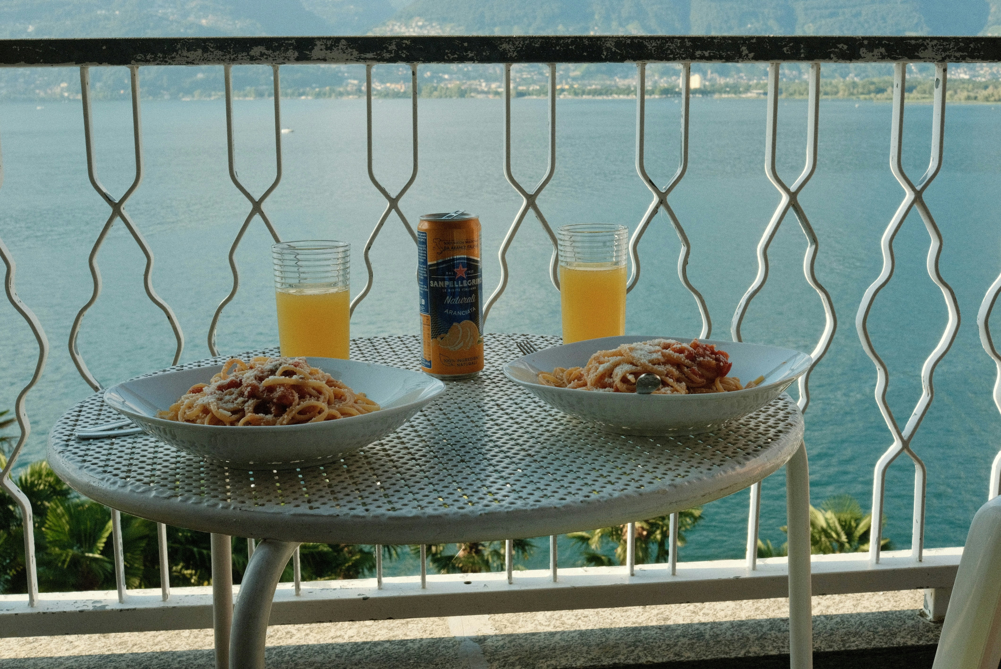 Enjoying the Dolce Vita at Lago Maggiore: With a breathtaking view of the tranquil lake, savor a delightful meal on the balcony. Plates of delicious pasta, paired with refreshing orange soda, are set on the table. The serene water and picturesque scenery provide a perfect backdrop for a leisurely dining experience. Here, life's simple pleasures are truly appreciated, and everyday stress feels miles away.