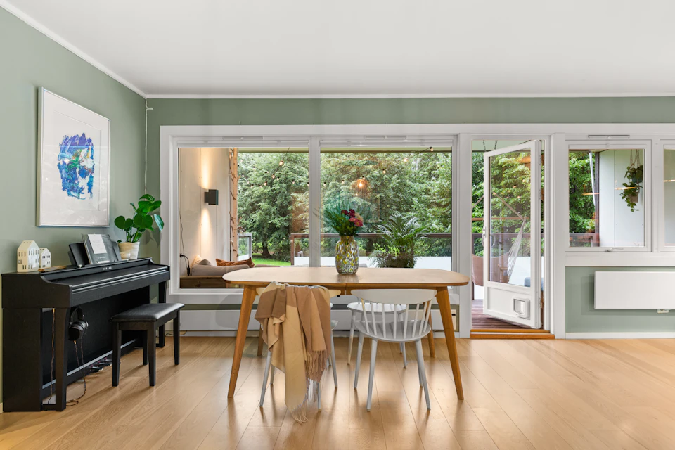 The image depicts a modern living room with a large window that offers a view of trees outside, allowing natural light to flood the space. The room features a wooden floor and white walls, creating a warm and inviting atmosphere. A black piano is positioned against one wall, suggesting musical interests or performances in this area. In front of the piano, there are two chairs - one white and one brown - arranged for seating. On the table near these chairs, there is a vase filled with flowers, adding a touch of elegance to the room. The overall design and layout of the living room create an open and airy feel, while still maintaining a sense of comfort and functionality.