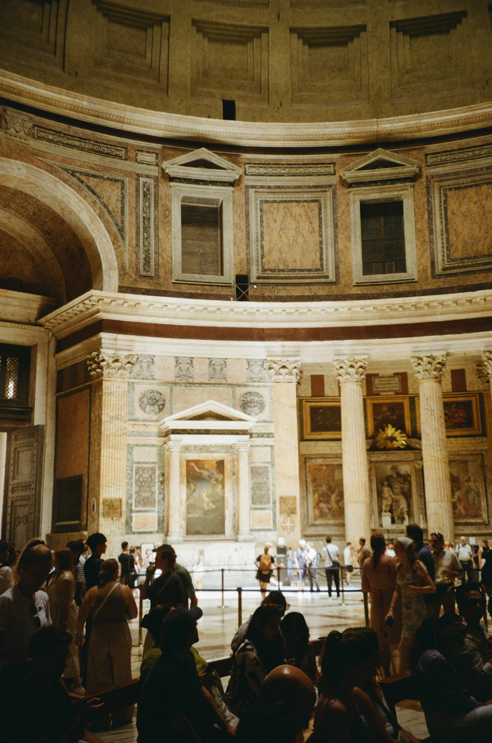 The image depicts a large, ornate room with a high ceiling and intricate architectural details. The walls are adorned with paintings and sculptures, adding to the grandeur of the space. A group of people can be seen gathered around the room, some standing in front of the windows while others are seated on benches or chairs scattered throughout the area.

The perspective of the image is from a low angle, looking up at the ceiling, which enhances the sense of awe and admiration for the architectural marvels present. The lighting in the room casts long shadows and highlights the intricate details of the walls and sculptures, creating a dramatic effect that emphasizes the grandeur of the space.