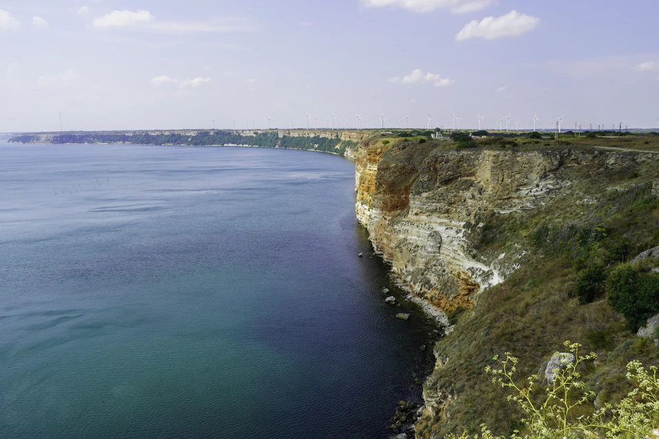 The image depicts a cliff overlooking a deep blue ocean, with a white cliff on one side and a green cliff on the other. The sky above is clear and blue, dotted with fluffy white clouds. In the distance, there are buildings visible along the shoreline, suggesting that this location may be near a populated area or coastal city.