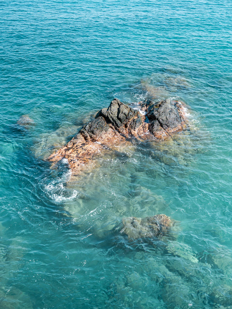 The image depicts a serene underwater scene with a large rock formation nestled amidst the turquoise waters. The rock, composed of various shades of brown and gray, is situated on the right side of the frame, surrounded by small waves that gently lap against its surface. The water surrounding the rock is a light blue color, contrasting beautifully with the darker hues of the rock. In the background, slightly to the left, there are more rocks visible, adding depth to the scene and creating a sense of scale.