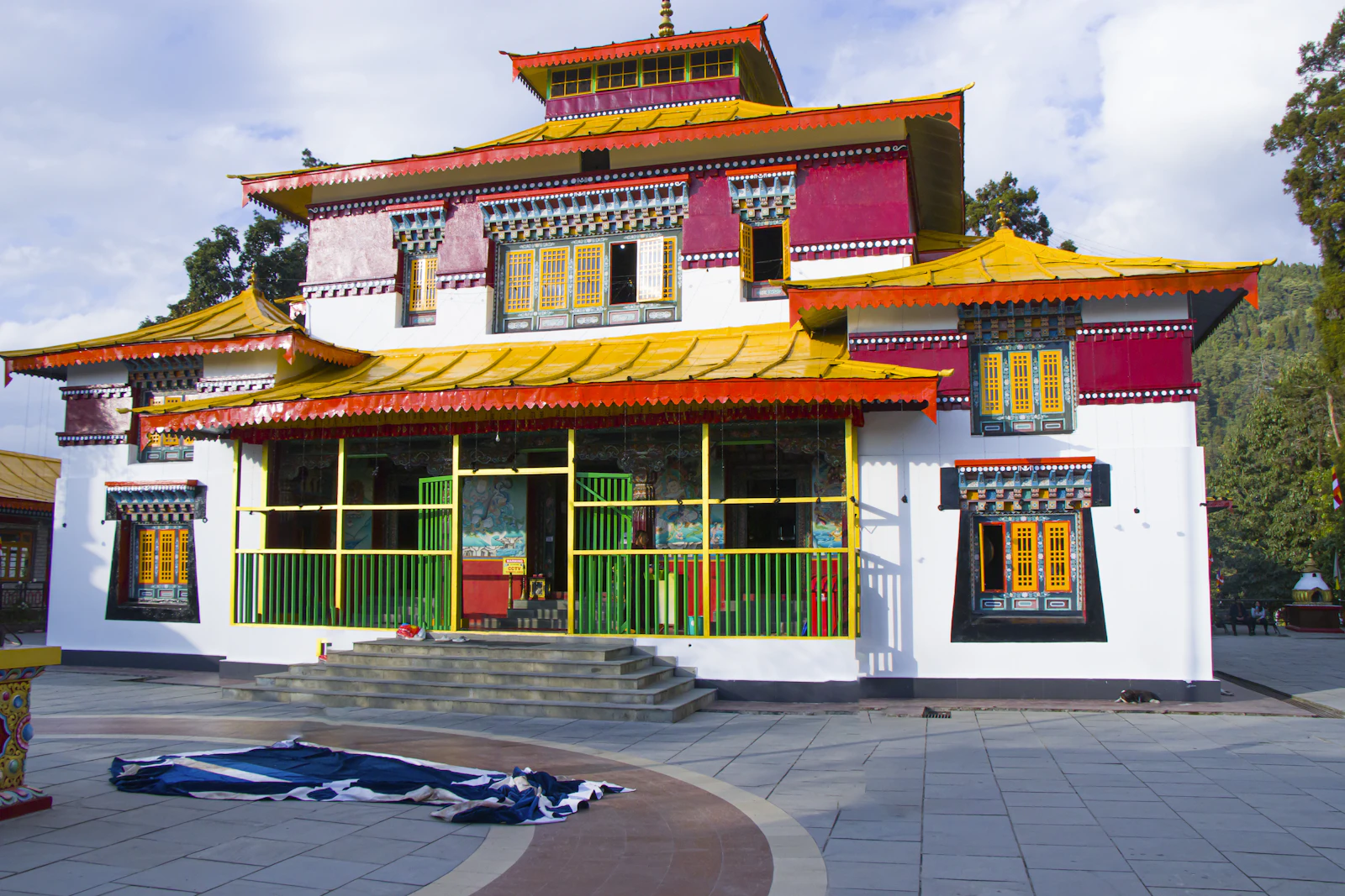A monastery temple in Pelling with tiered yellow roofs and painted windows above a paved courtyard, forest behind