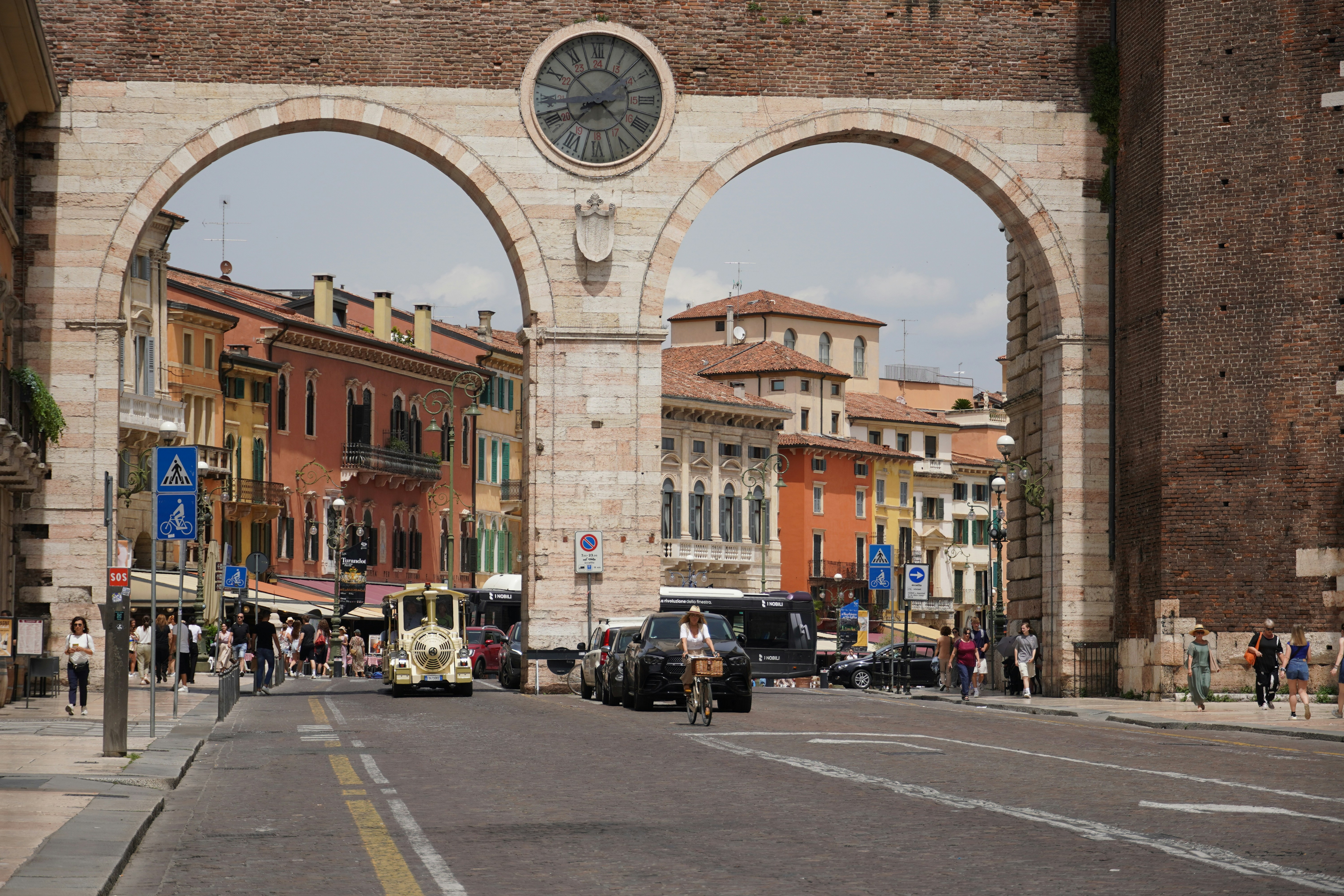 Verona - Historic archway with a clock against a vibrant street, with colorful buildings, people, and cars in the background.