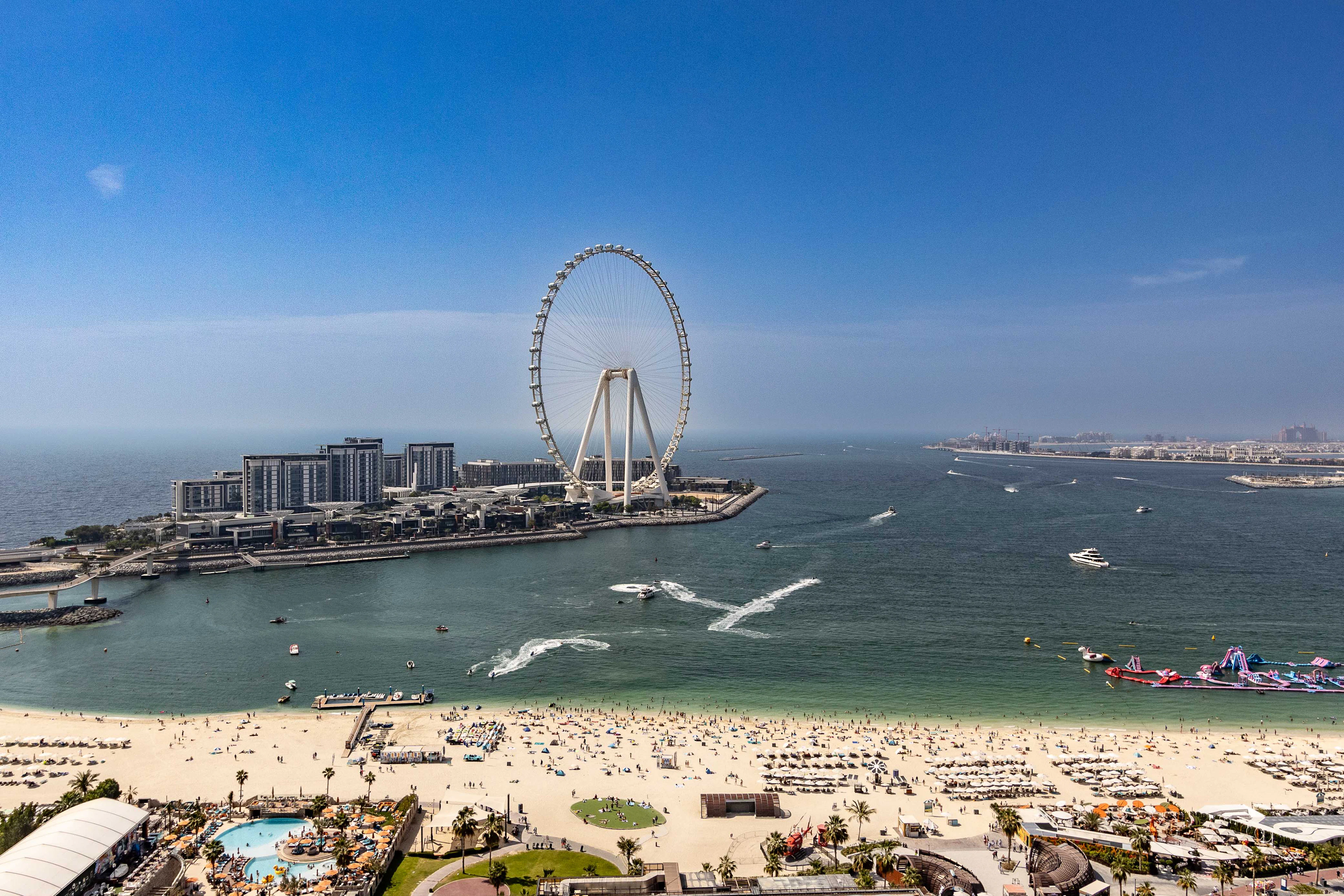JBR beach from a high balcony on a clear day: a broad crescent of pale sand crowded with sunshades, green-blue water crossed by the wakes of jet skis, and the white Ain Dubai observation wheel standing over Bluewaters Island opposite