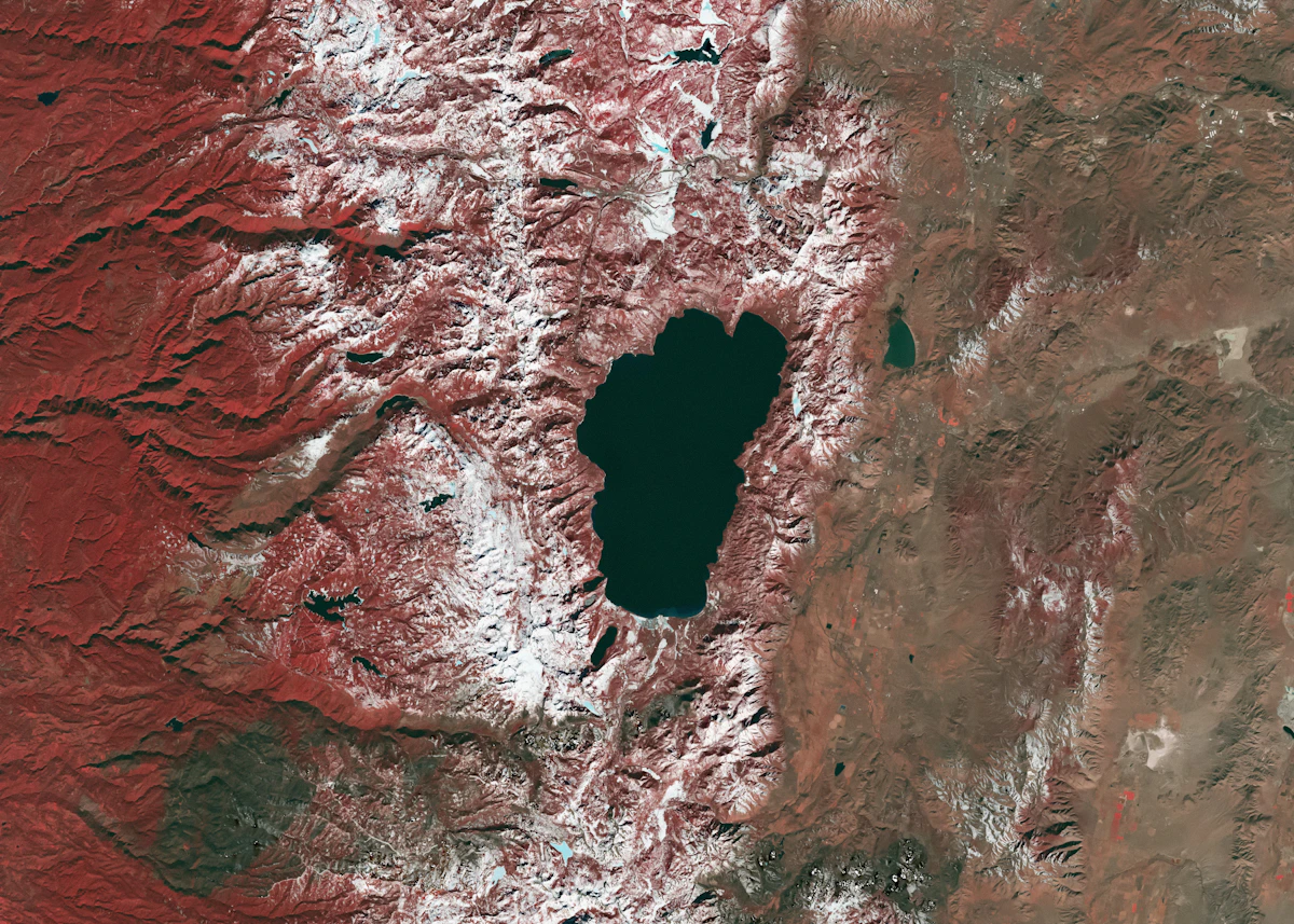 A satellite view of a dark lake surrounded by mountains, showing broad shapes clearly but withholding finer ground-level detail until the view is zoomed in, analogous to a coarse memory summary that a model can request higher resolution from on demand