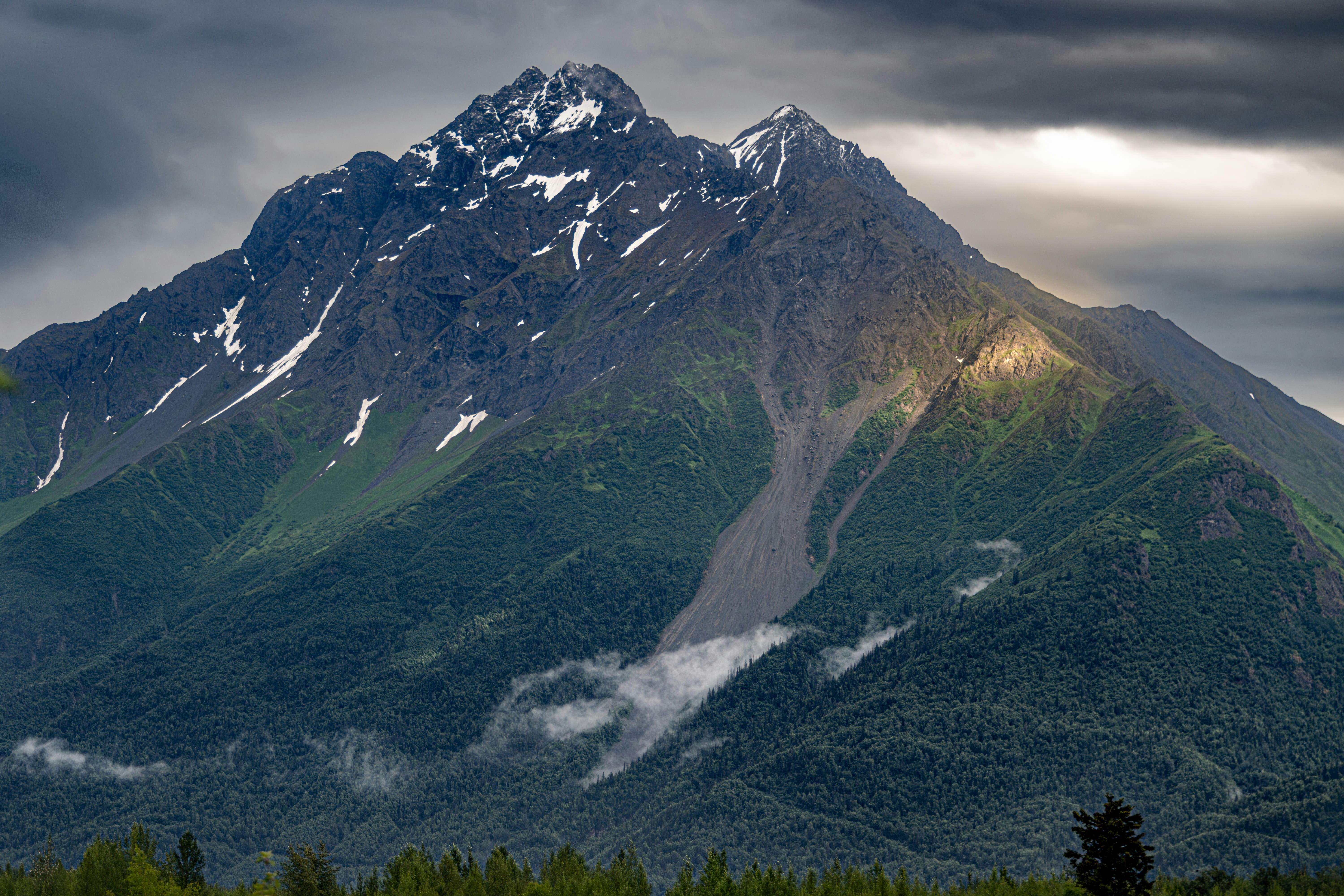 A large mountain covered in snow under a cloudy sky, A stunning Alaskan mountain landscape showcasing vibrant green valleys and dramatic, cloud-covered peaks.