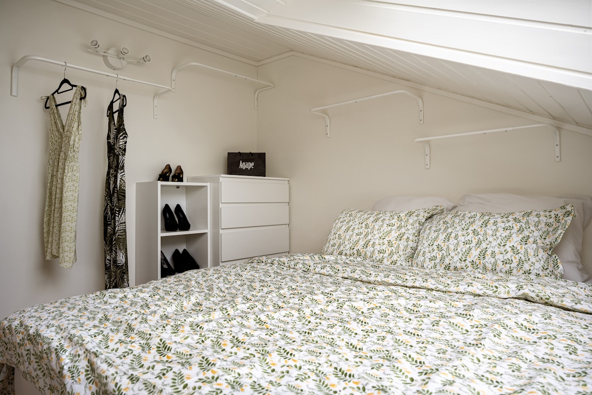 Small attic bedroom with wall-mounted clothes rack, compact white dresser, and shoe storage shelf for creative space-saving storage
