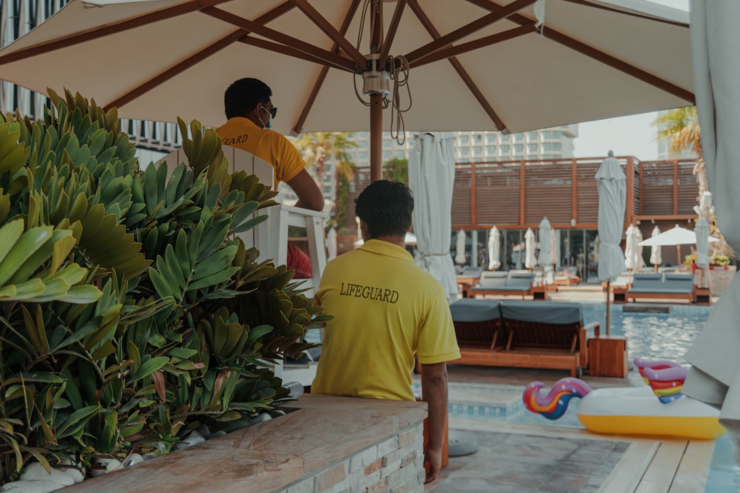 A man standing under an umbrella next to a pool, Lifeguards on duty by the poolside in a modern resort setting
