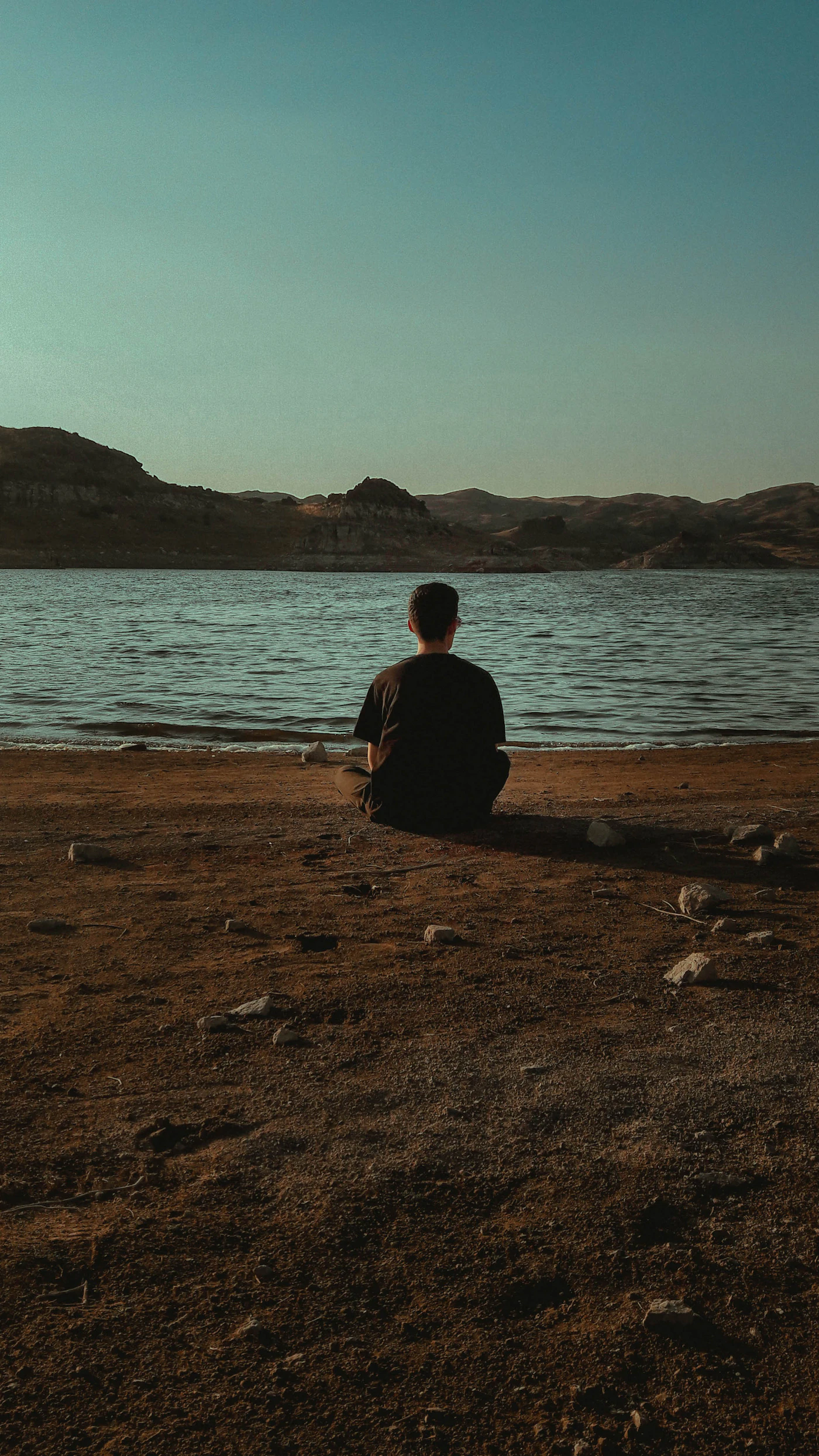 A man sits alone by a still lake, back to camera, mountains in the distance -- a moment of solitary stillness