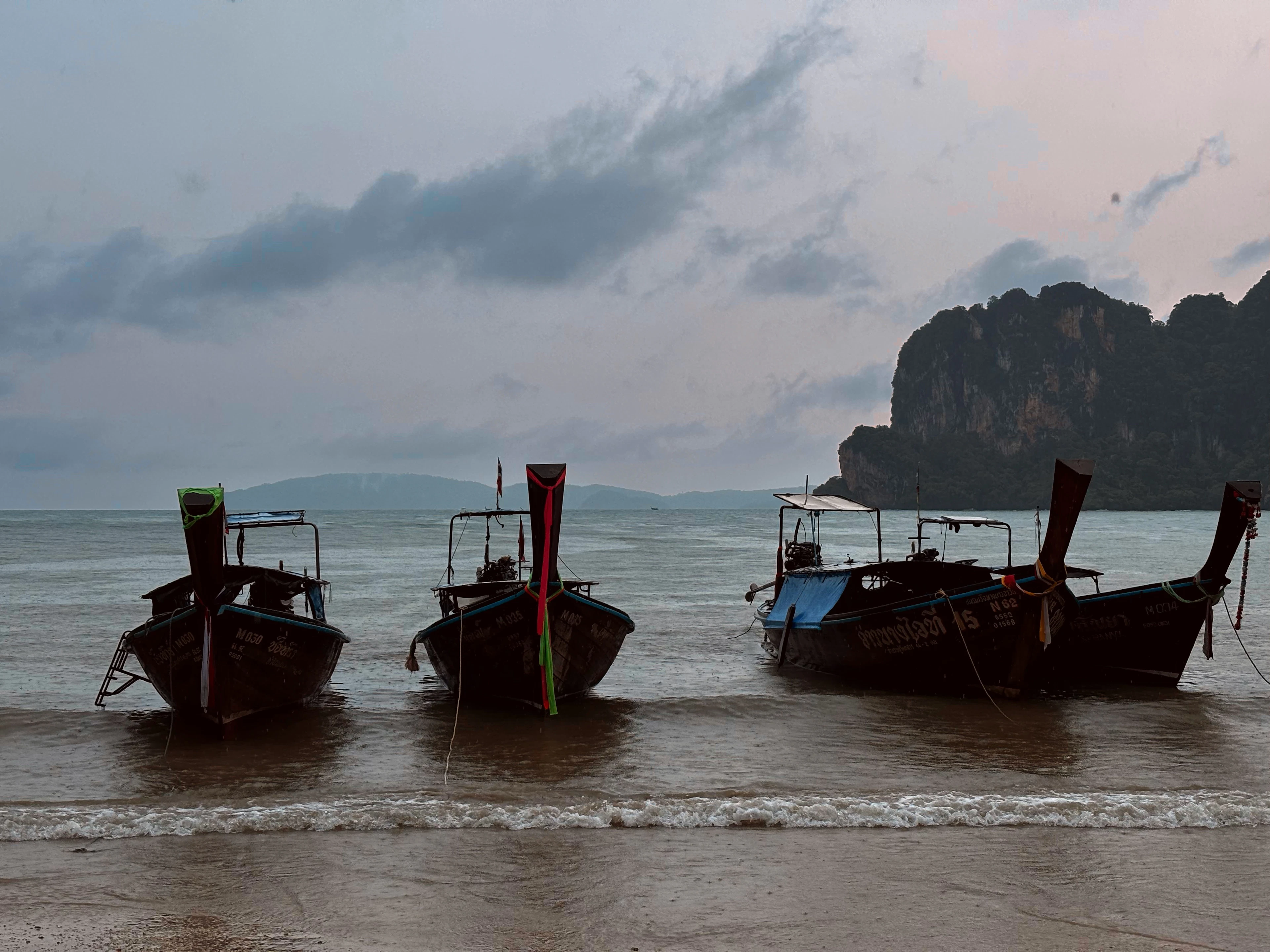 Three wooden longtail boats moored on a Krabi beach beneath a moody, cloud-streaked dusk sky.