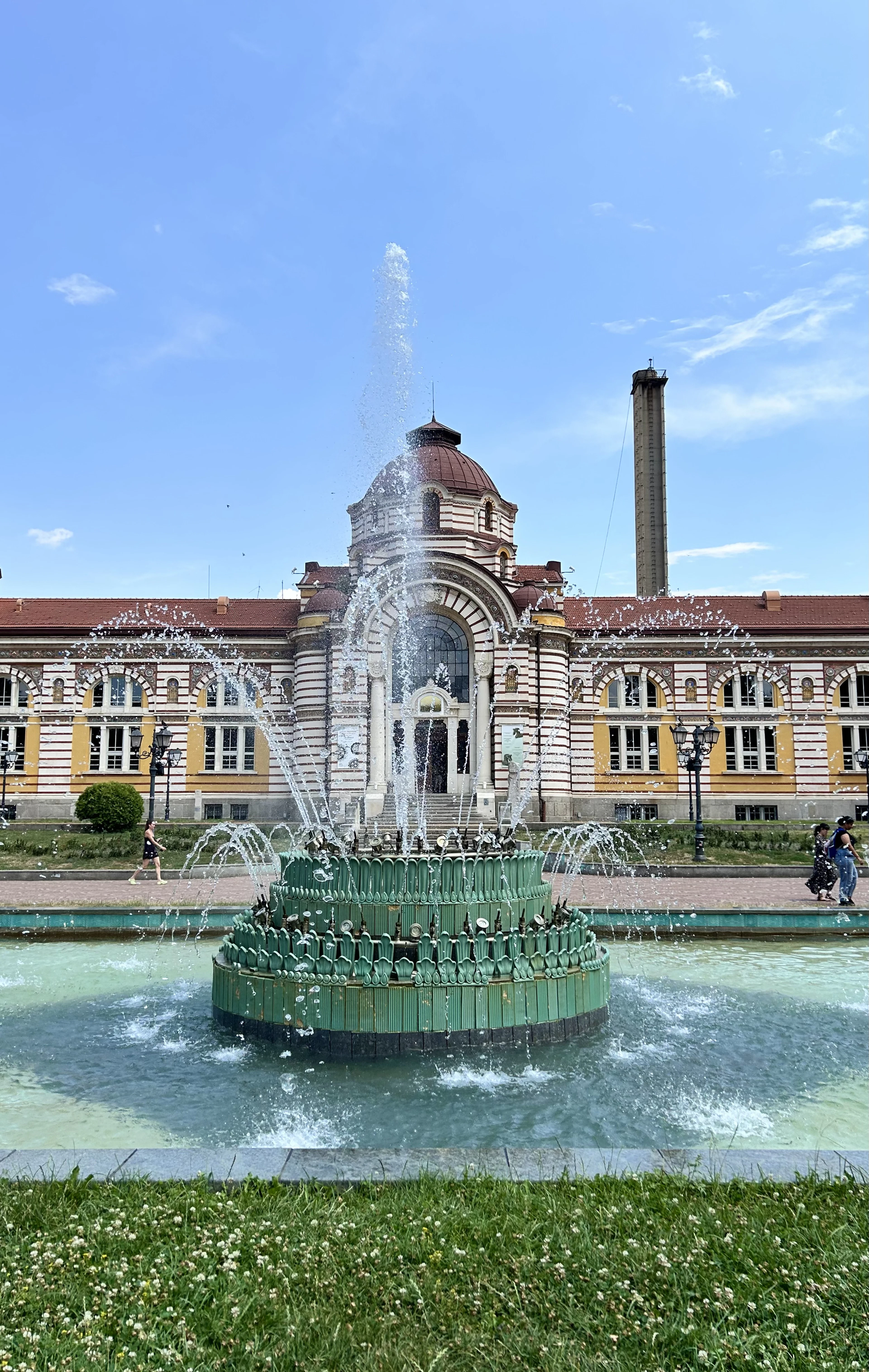 The ochre and red-brick facade of Sofia's Central Mineral Baths rising behind its green-tiled fountain, jets of water arcing into the pool under a clear blue sky.