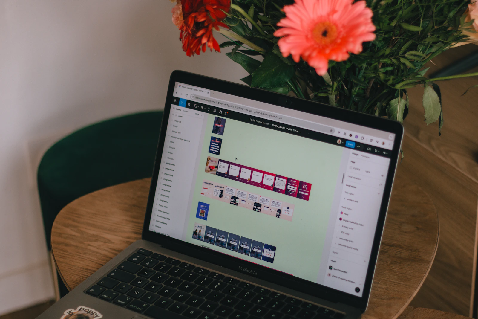 A laptop open to a design canvas with several screen layouts, next to coral gerbera flowers on a wooden table