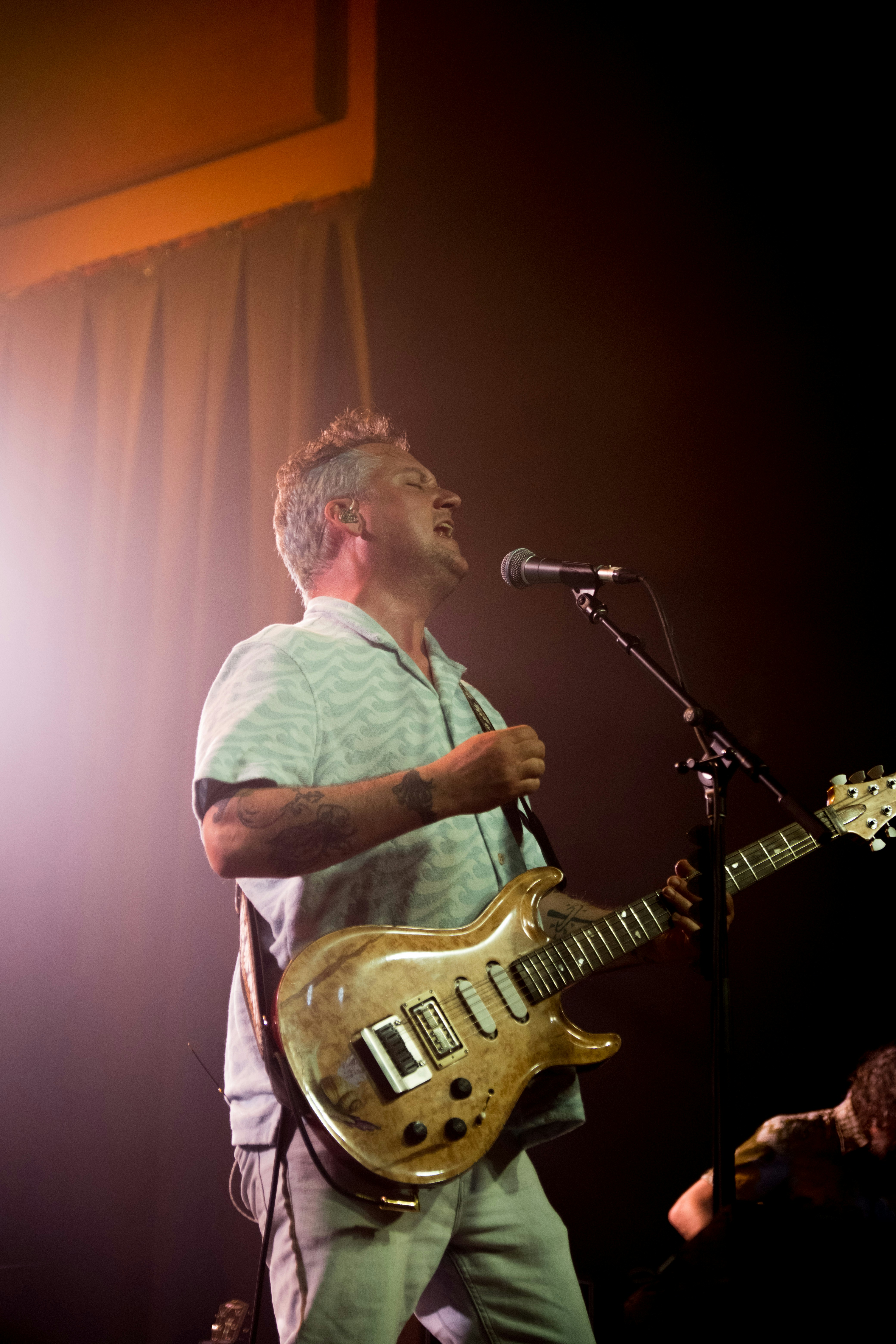 Cantante masculino actuando en el escenario con guitarra, fondo tropical, iluminación cálida del escenario