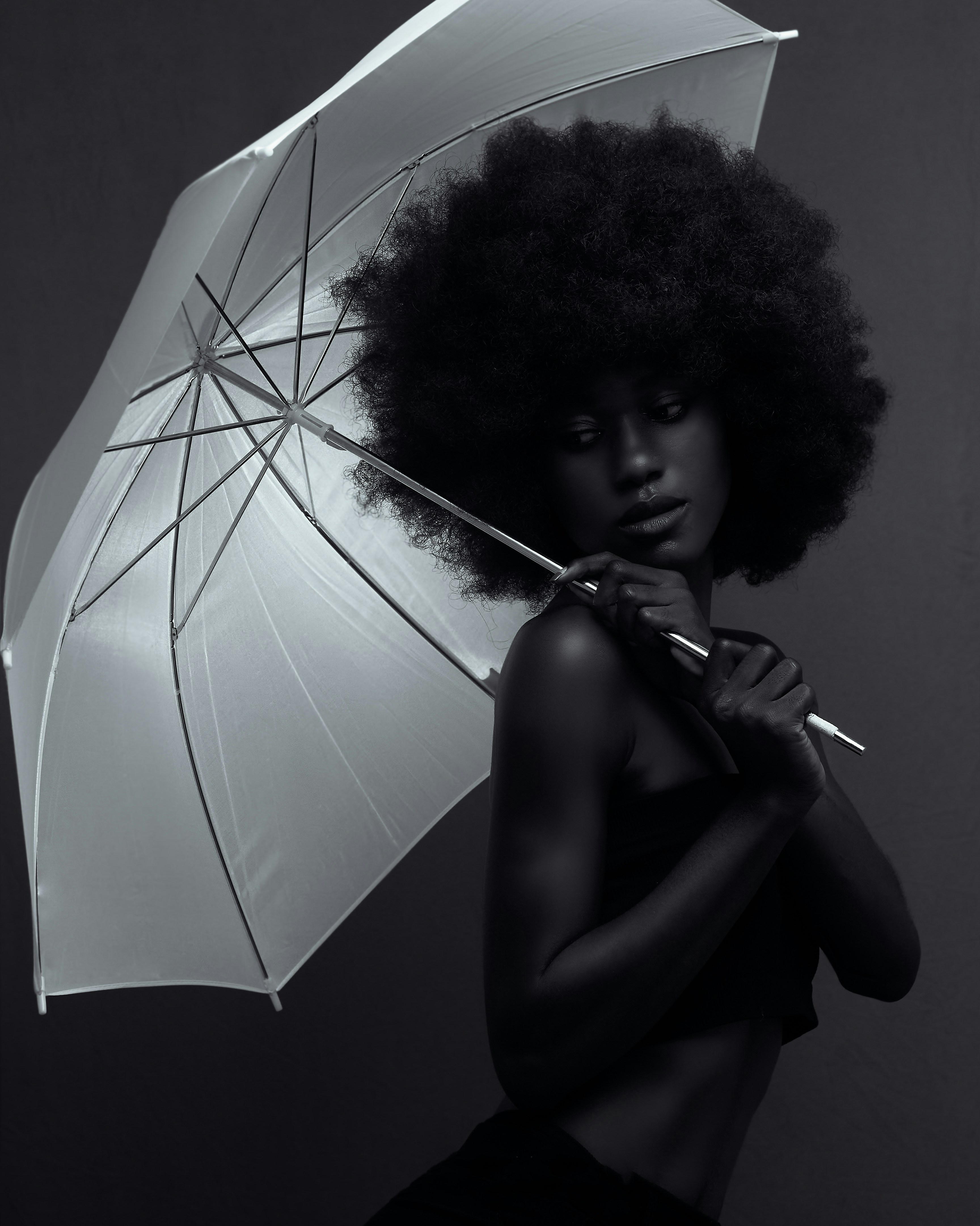 A black and white portrait of a Black woman with an afro hairstyle, posing while holding a large white umbrella over her shoulder. 