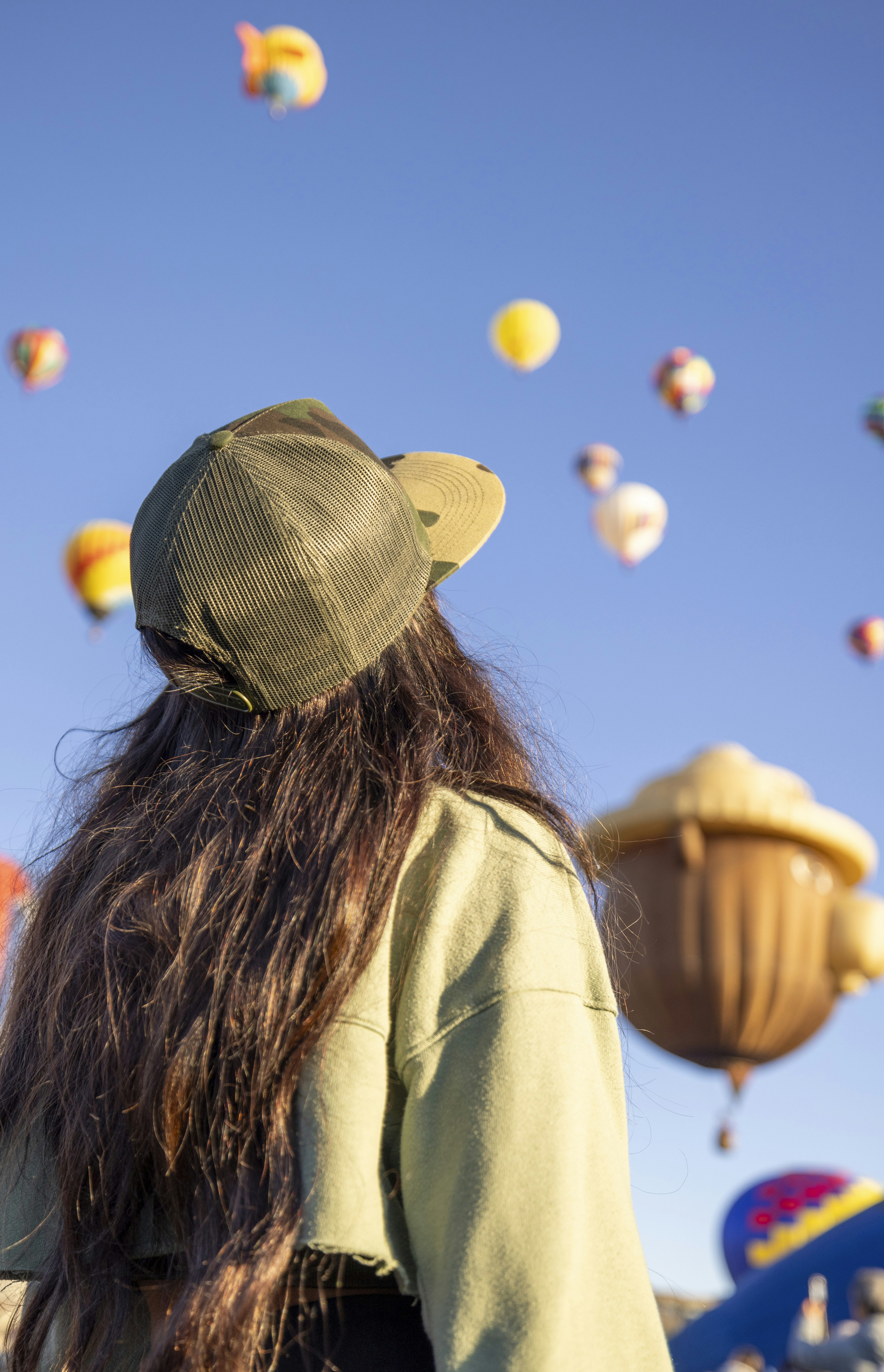 Candid photograph of a person with long hair in a green cap, seen from behind, as vibrant balloons drift across a clear blue sky.