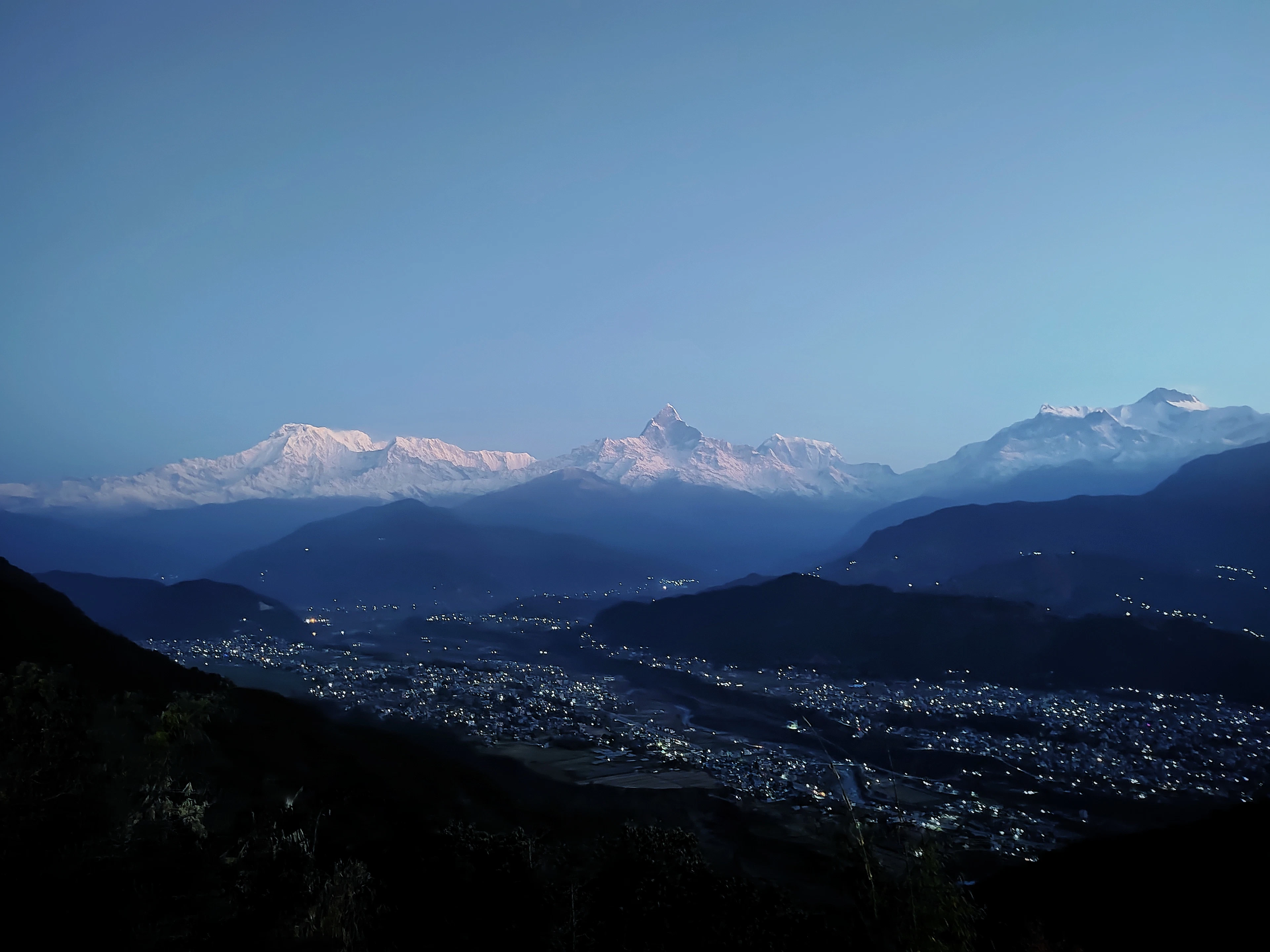 The lights of Pokhara scattered across the dark valley before sunrise, seen from Sarangkot, with the snow-covered Annapurna peaks catching the first pale light above.