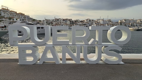 A metal sign that says puerto banus in front of a body of water