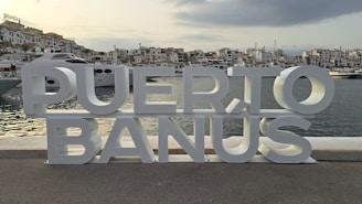 A metal sign that says puerto banus in front of a body of water