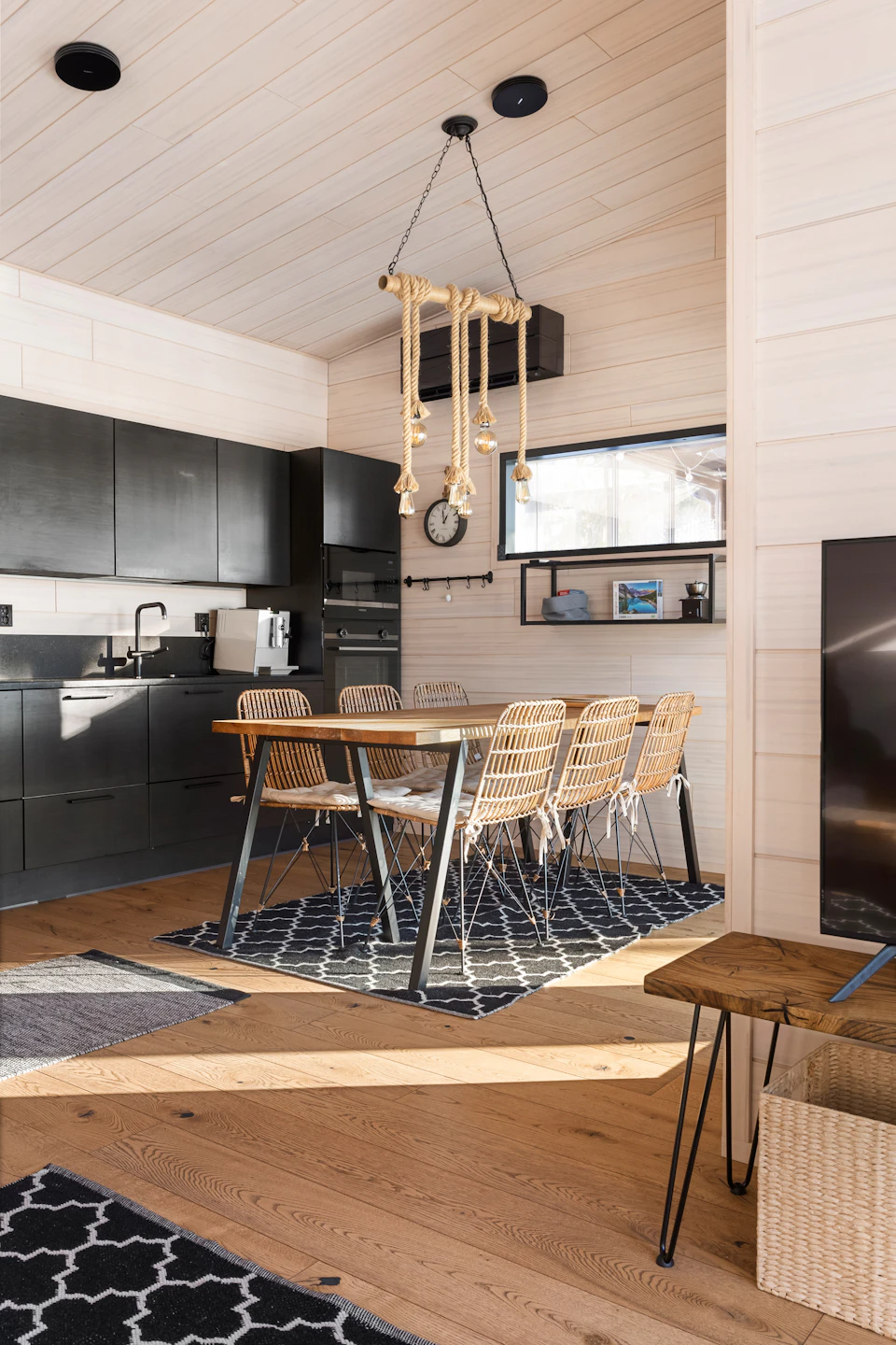 The image depicts a modern kitchen and dining area with white walls, wooden floors, and black cabinets. The kitchen features a sink on one side and a window above it, allowing natural light to enter the space. A chandelier hangs from the ceiling, adding an elegant touch to the room. In the center of the room is a large table surrounded by six chairs, suggesting a dining area for family meals or gatherings.

On the right side of the image, there is a television mounted on the wall, and a small wooden shelf holds various items such as bottles and other kitchenware. The floor has a black and white geometric pattern rug in front of the table, adding visual interest to the space.