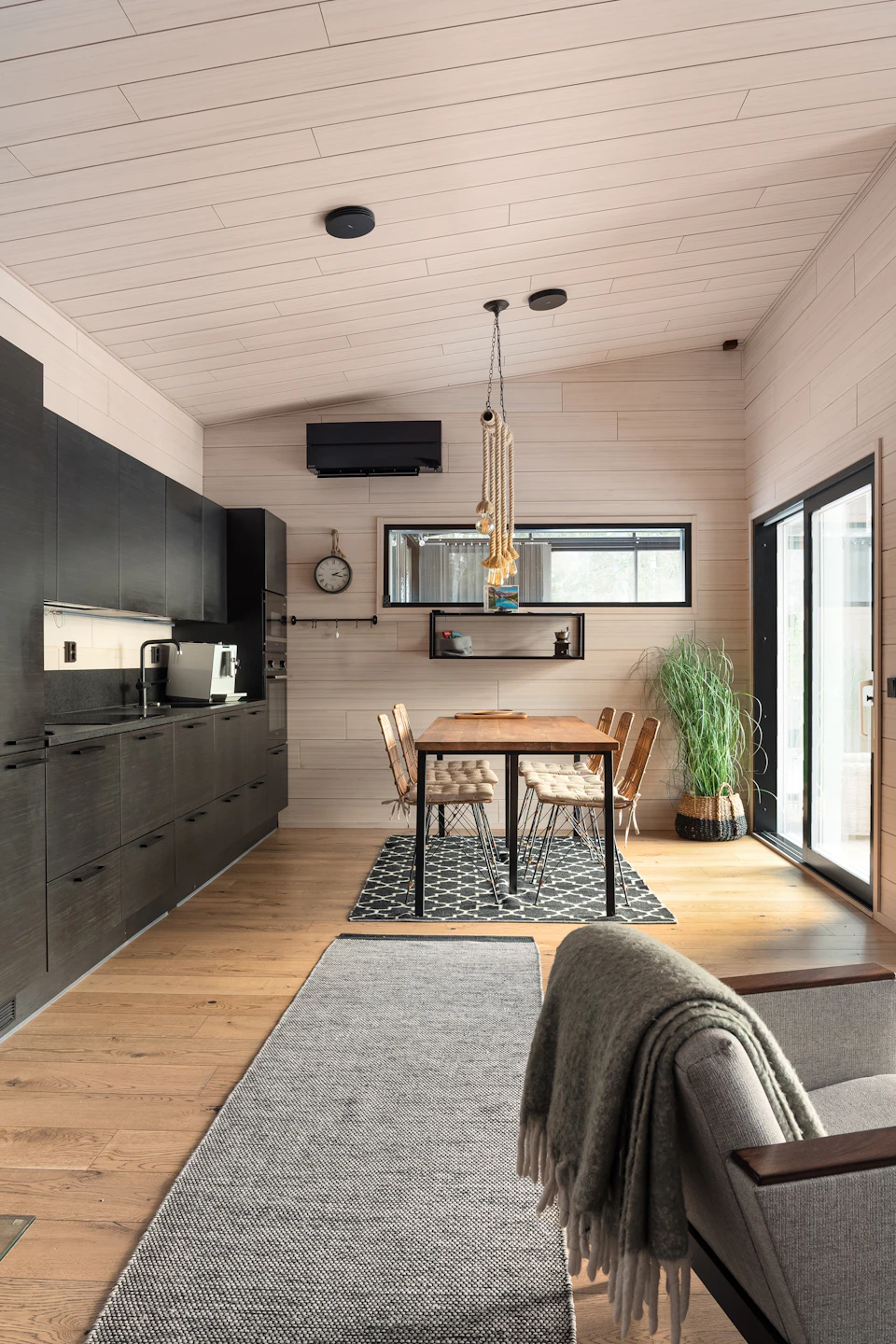 The image depicts a modern kitchen and dining area with white walls, wooden floors, and black cabinets. The kitchen features a sink on one side and a refrigerator on another. A window above the sink allows natural light to enter the space. In the center of the room is a dining table surrounded by six chairs, two of which are empty. On the table, there is a vase with flowers and a clock. A gray rug covers the floor in front of the table. The walls are painted white, creating a bright and airy atmosphere.
