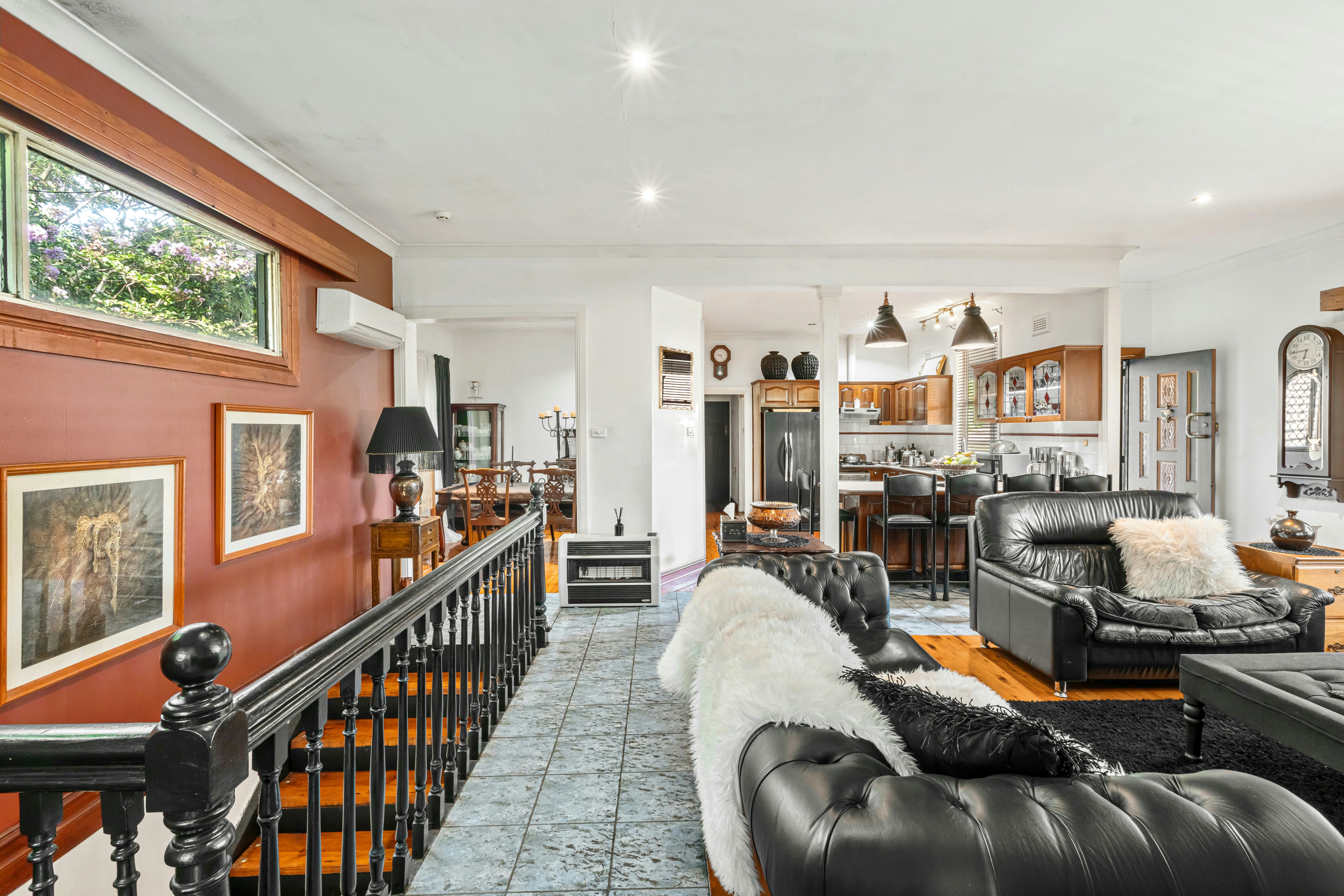 An eclectic home interior features black leather sofas, a dark staircase, reddish-brown walls, and views into a traditional dining room and kitchen.