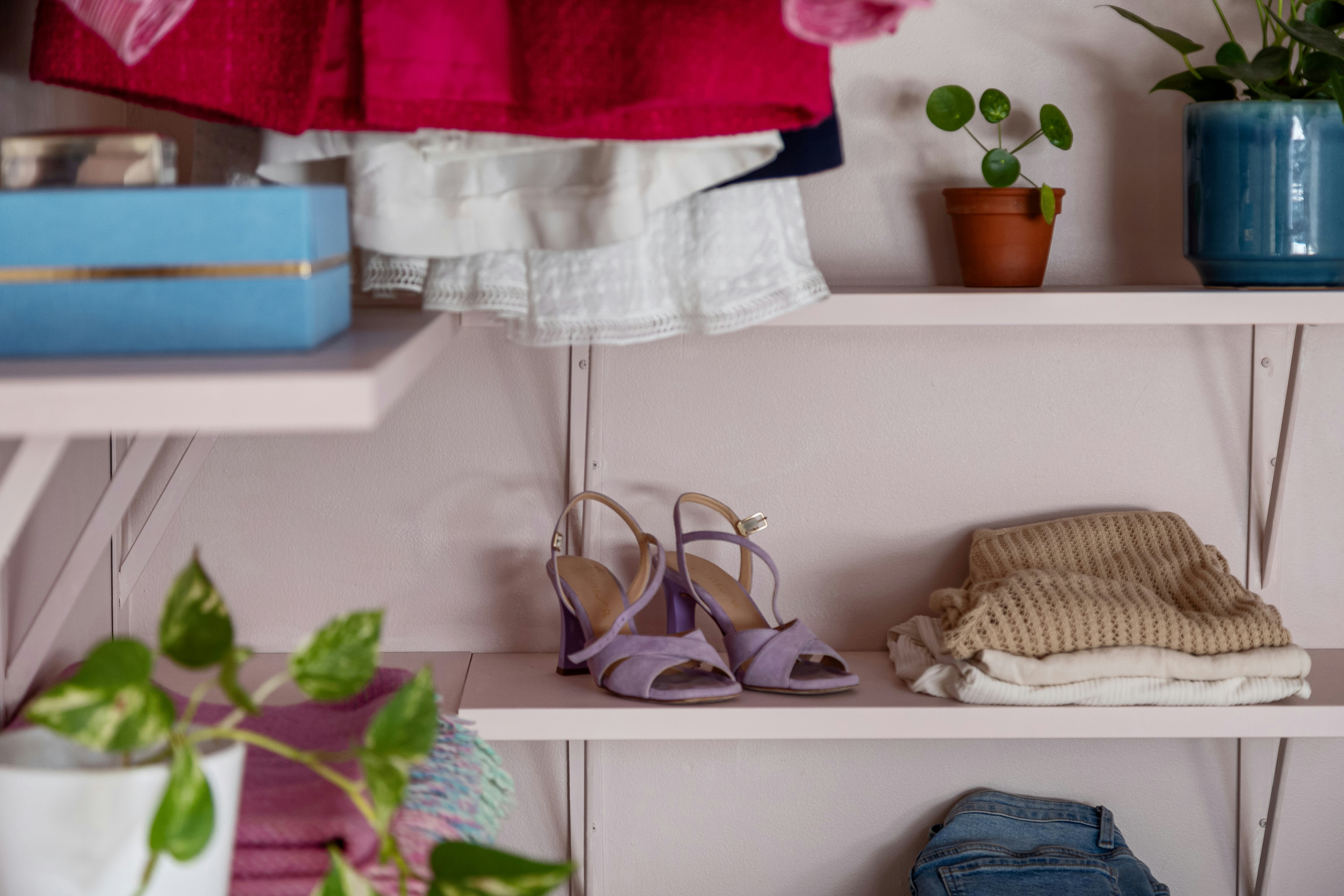 Open wardrobe with pink shelves holding lavender heeled sandals, folded sweaters, hanging colorful clothes, and small potted plants