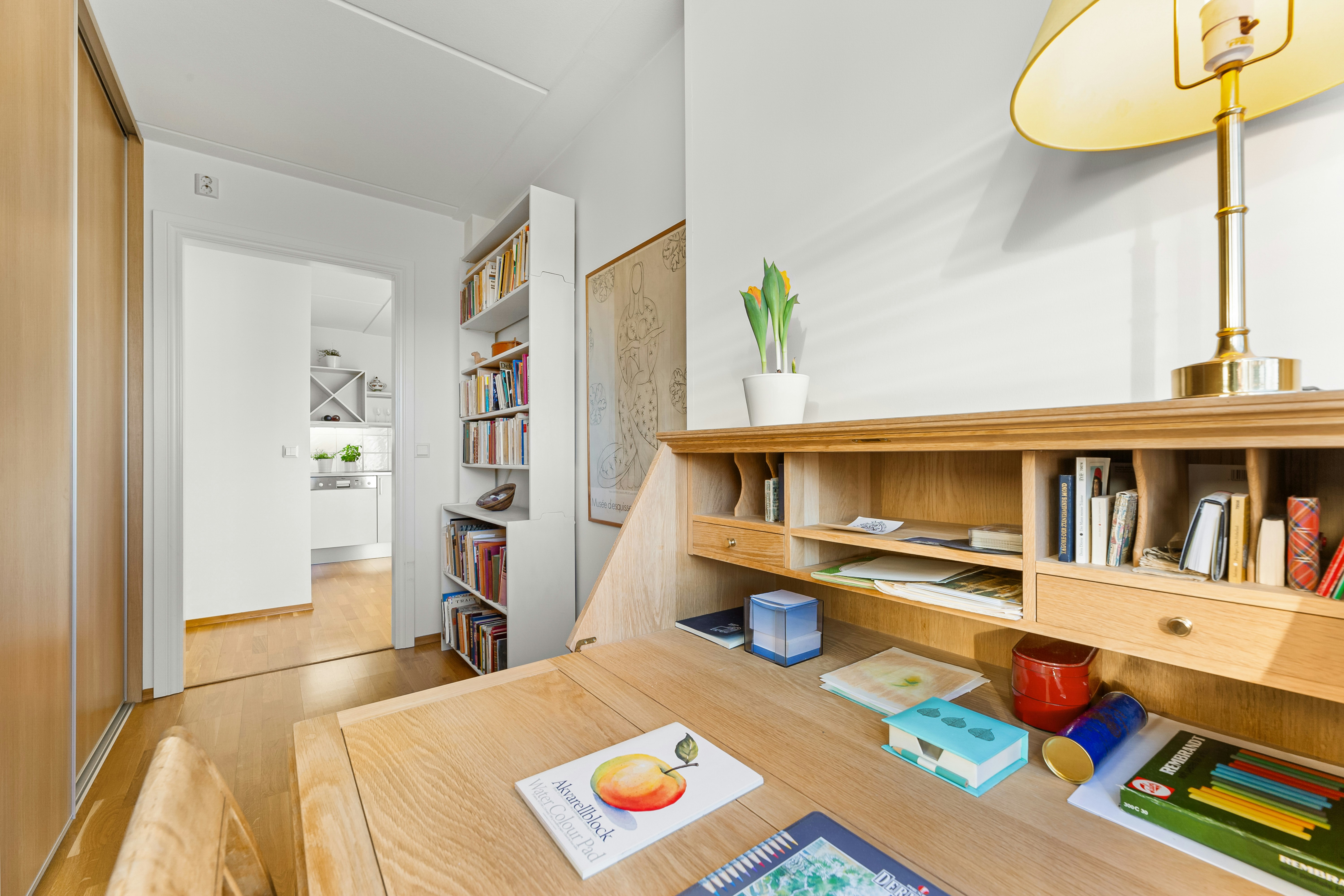 A room with a desk, bookshelf and a lamp