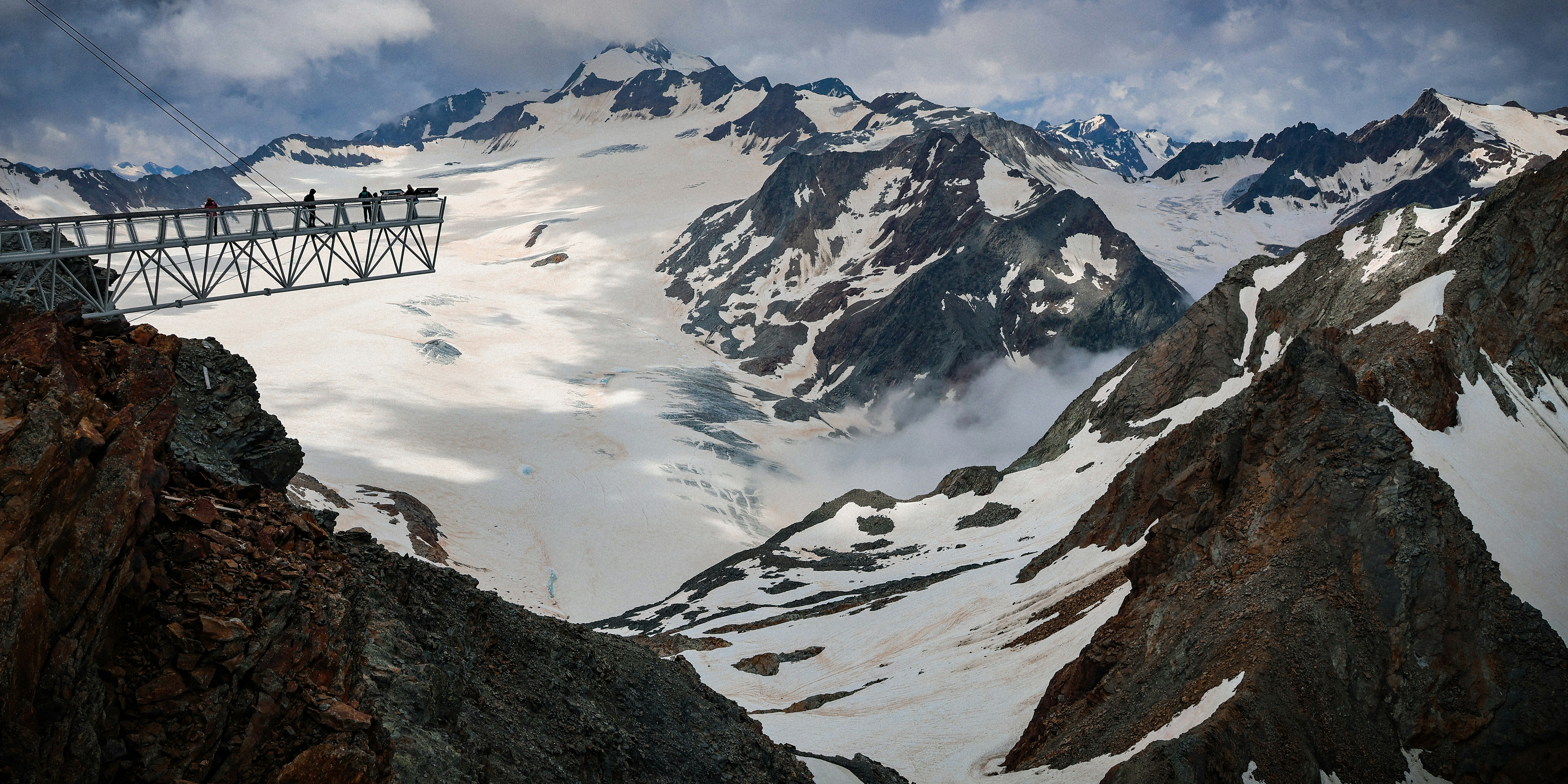 Landscape photograph featuring jagged snow-capped peaks, a glacier valley, and a metal viewing structure on the left beneath a cloudy sky.