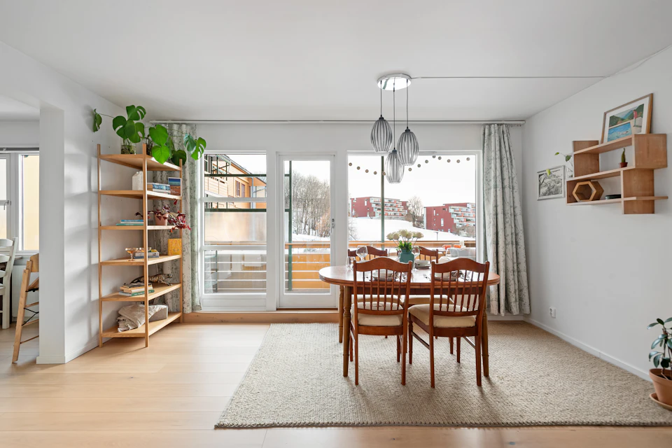 The image depicts a modern, open-concept living room with white walls and a light wood floor. The room features a large window that offers a view of trees outside, allowing natural light to flood the space. A wooden bookshelf stands against one wall, filled with various items such as plants and decorative objects.

In the center of the room is a dining table surrounded by four chairs, two on each side. The table is set for three people, suggesting it may be used for social gatherings or family meals. A rug covers the floor in front of the table, adding warmth to the space.

On one wall, there is a door leading outside, and another wall features a shelf with more plants and decorative items.