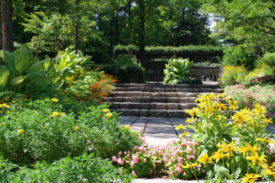 The image depicts a serene garden scene with a stone path winding through it, flanked by an array of colorful flowers and lush green plants. The path is bordered by a variety of plants, including yellow daisies, pink and white flowers, and orange marigolds. In the background, there are trees providing shade and adding depth to the landscape.
