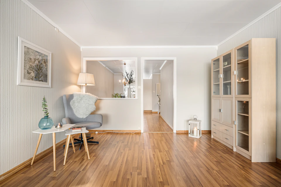 The image depicts a serene and well-lit living room with a large window that allows natural light to flood the space, illuminating the hardwood floor. The room features white walls and furniture, creating a clean and minimalist aesthetic. A gray armchair is positioned near a wooden coffee table, which holds a vase of flowers.

In one corner of the room, there's a bookshelf filled with various items, including several books that add an intellectual touch to the space. The hardwood floor extends throughout the room, enhancing its spaciousness and elegance. A doorway can be seen in the background, leading to another part of the house or possibly connecting two rooms together.