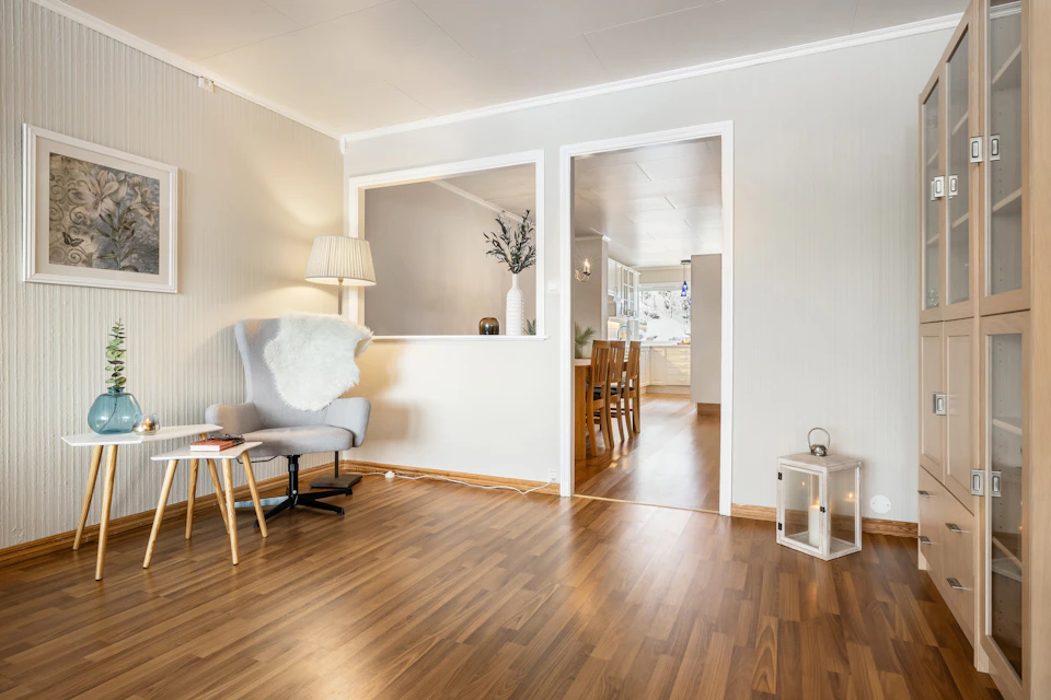 The image depicts a serene and well-lit living room with a large window on one side, allowing natural light to flood the space. The floor is made of hardwood, adding warmth and elegance to the room. A white armchair sits in the center, facing a wooden coffee table that holds a vase filled with flowers.

On either side of the room are two windows, each adorned with white curtains that allow for privacy while still letting in light. The walls are painted white, creating a bright and airy atmosphere. In one corner of the room, there is a bookshelf housing an array of books and decorative items. A doorway can be seen on the right side of the image, leading to another part of the house or possibly connecting with the outdoors.