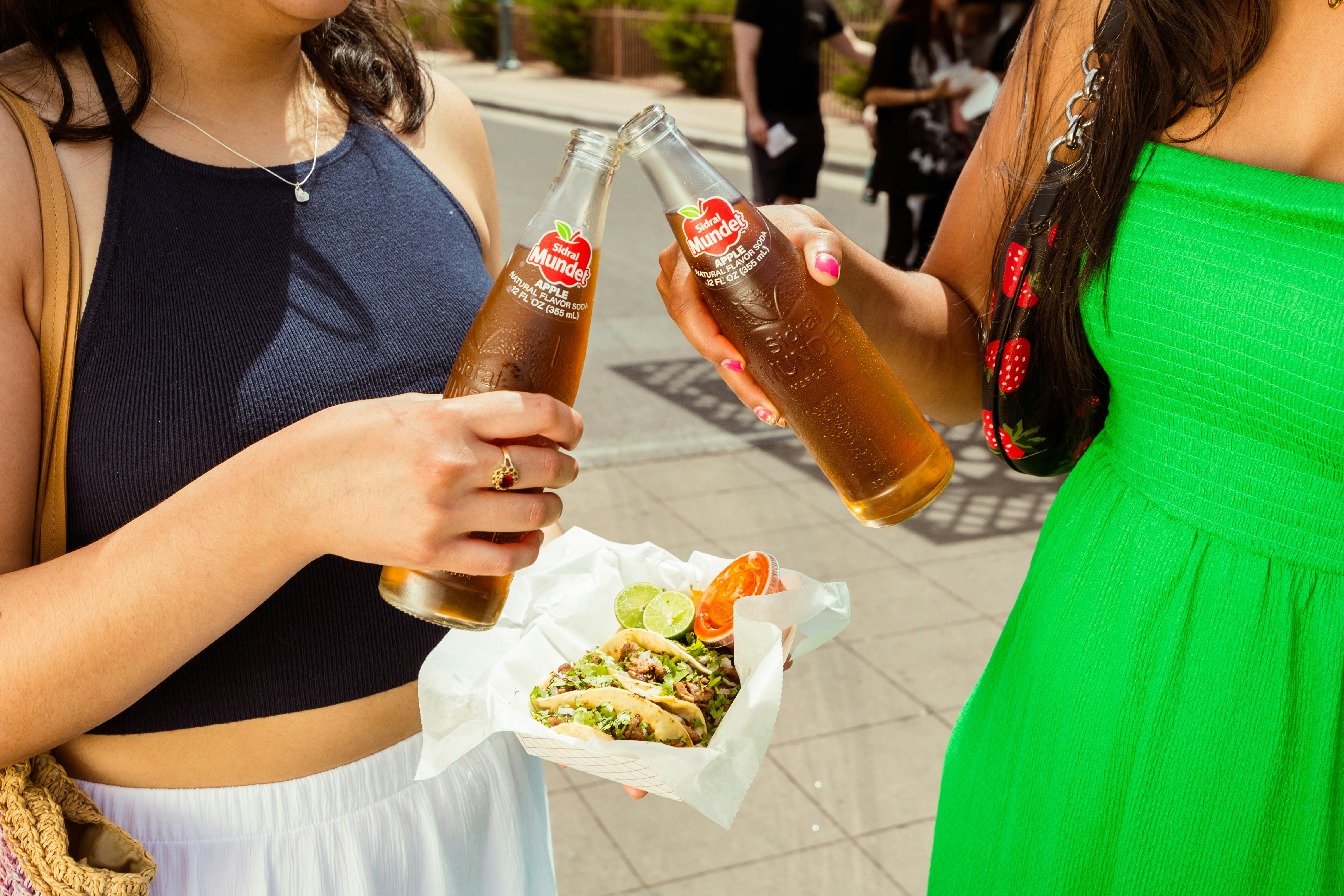 A couple of women standing next to each other, Two friends enjoying refreshing Sidral Mundet glass bottle sodas with their tacos from Luchador food truck.