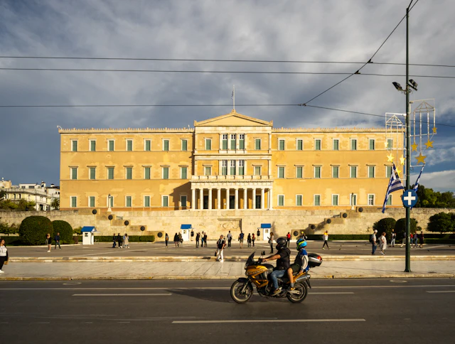 The Hellenic Parliament building in Syntagma, with a motorcycle passing on the street and Greek and EU flags flying.