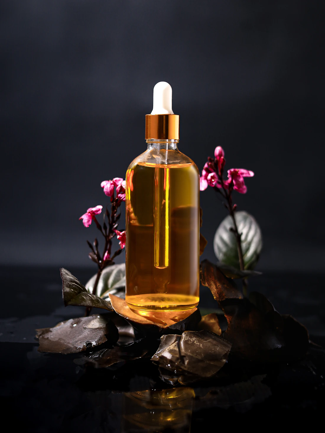 A glass bottle of golden facial oil with a copper dropper, set among dark stones and pink blossoms