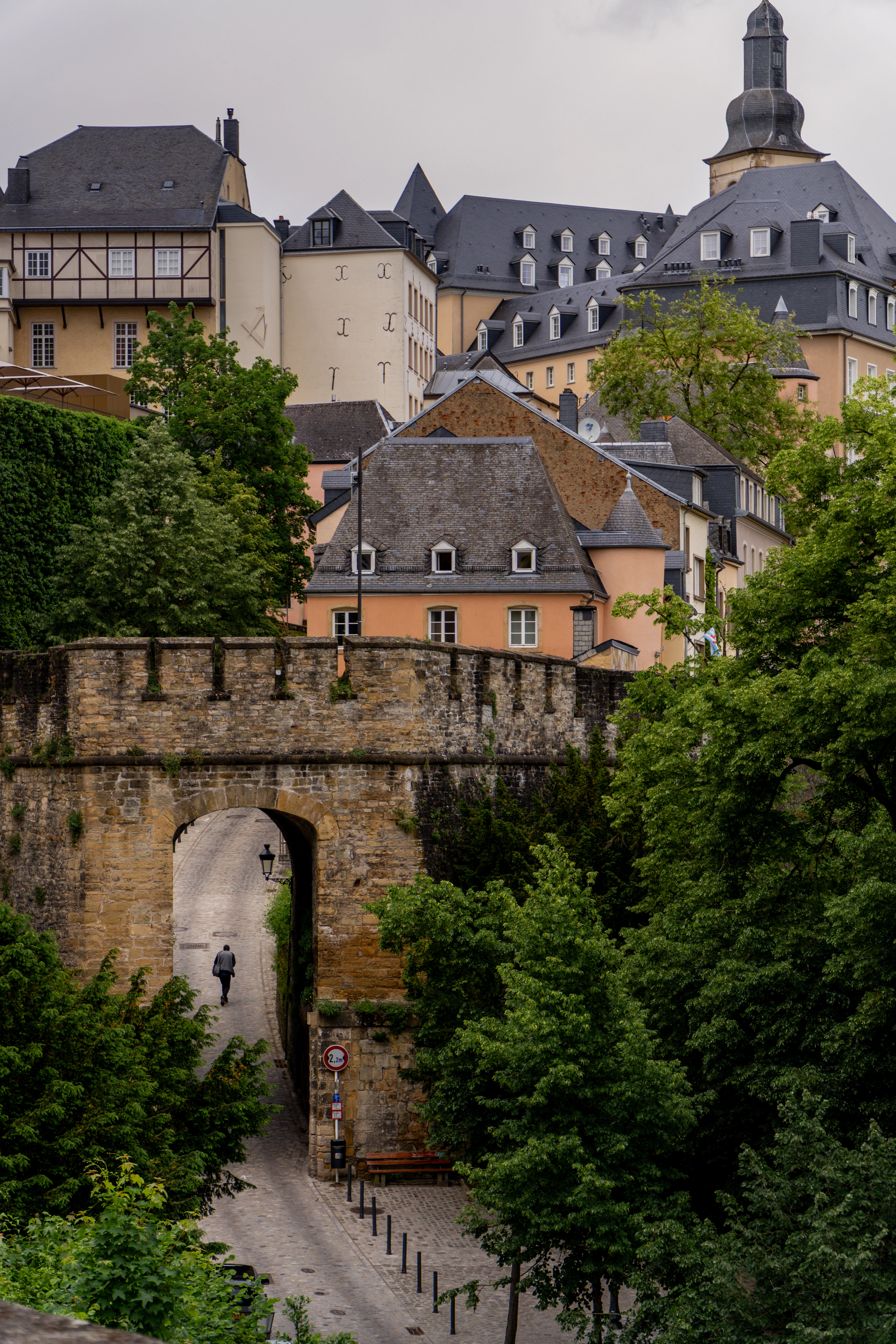 A person walks through a stone archway gate in an old fortress wall surrounded by lush green trees.