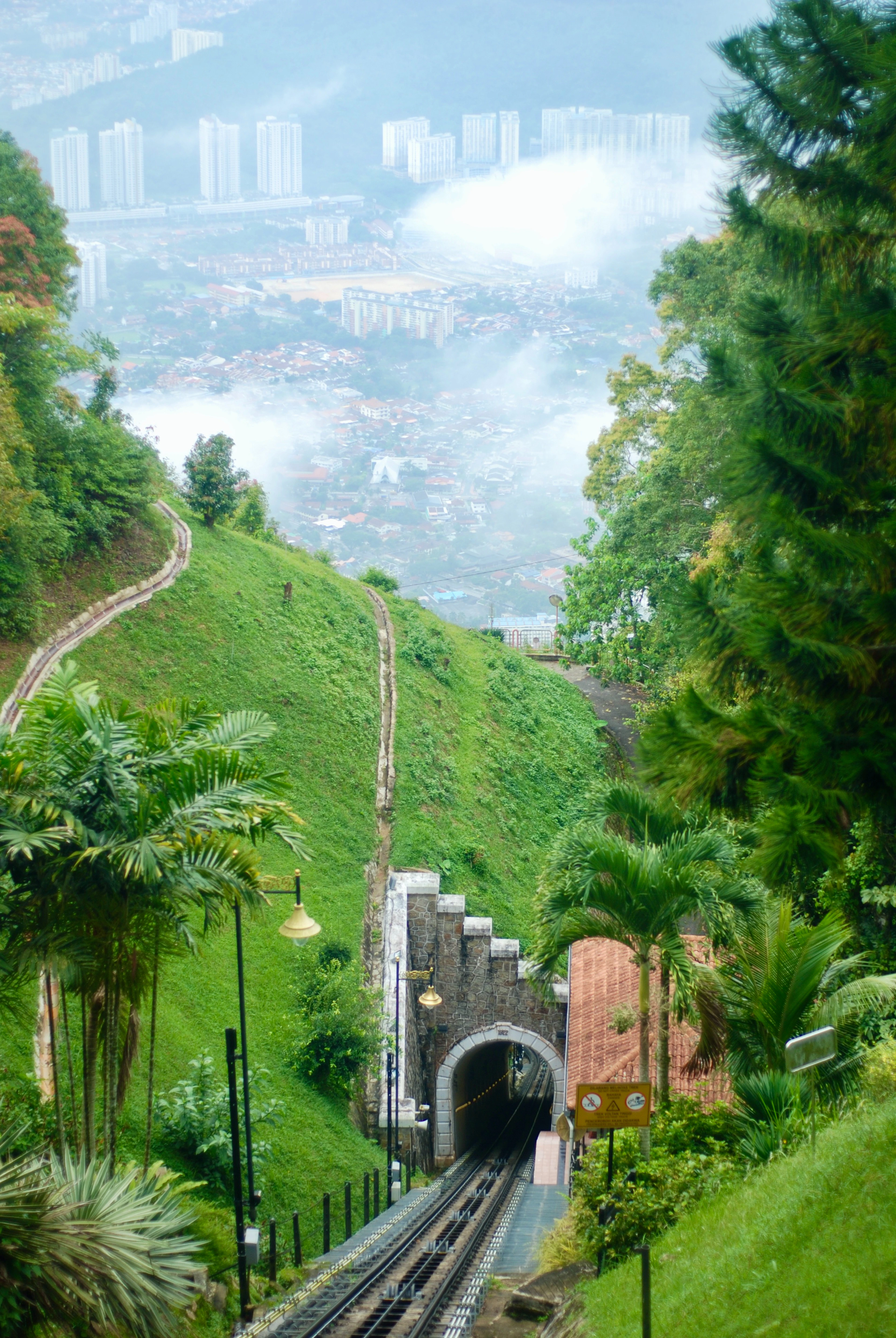 The Penang Hill funicular track dropping down a steep emerald hillside into its stone tunnel mouth, palms and lamp posts along the cutting and mist drifting over the rooftops of George Town far below.