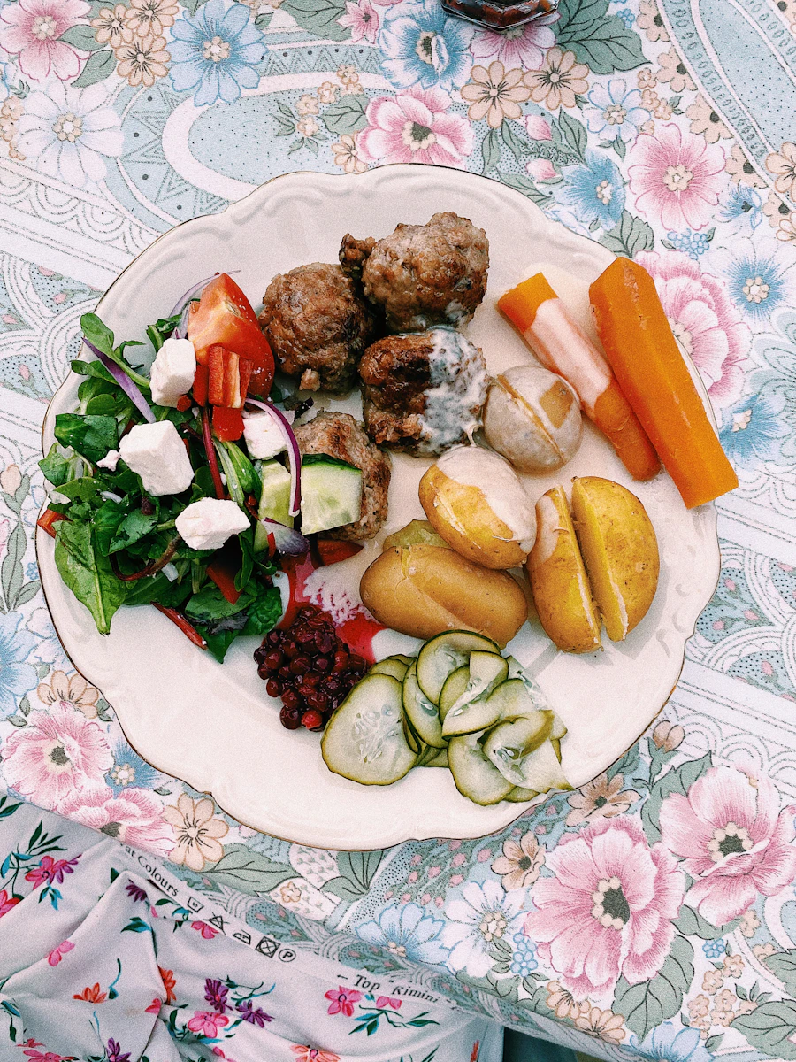 A plate of meatballs with boiled potatoes, pickled cucumber and vegetables on a floral tablecloth