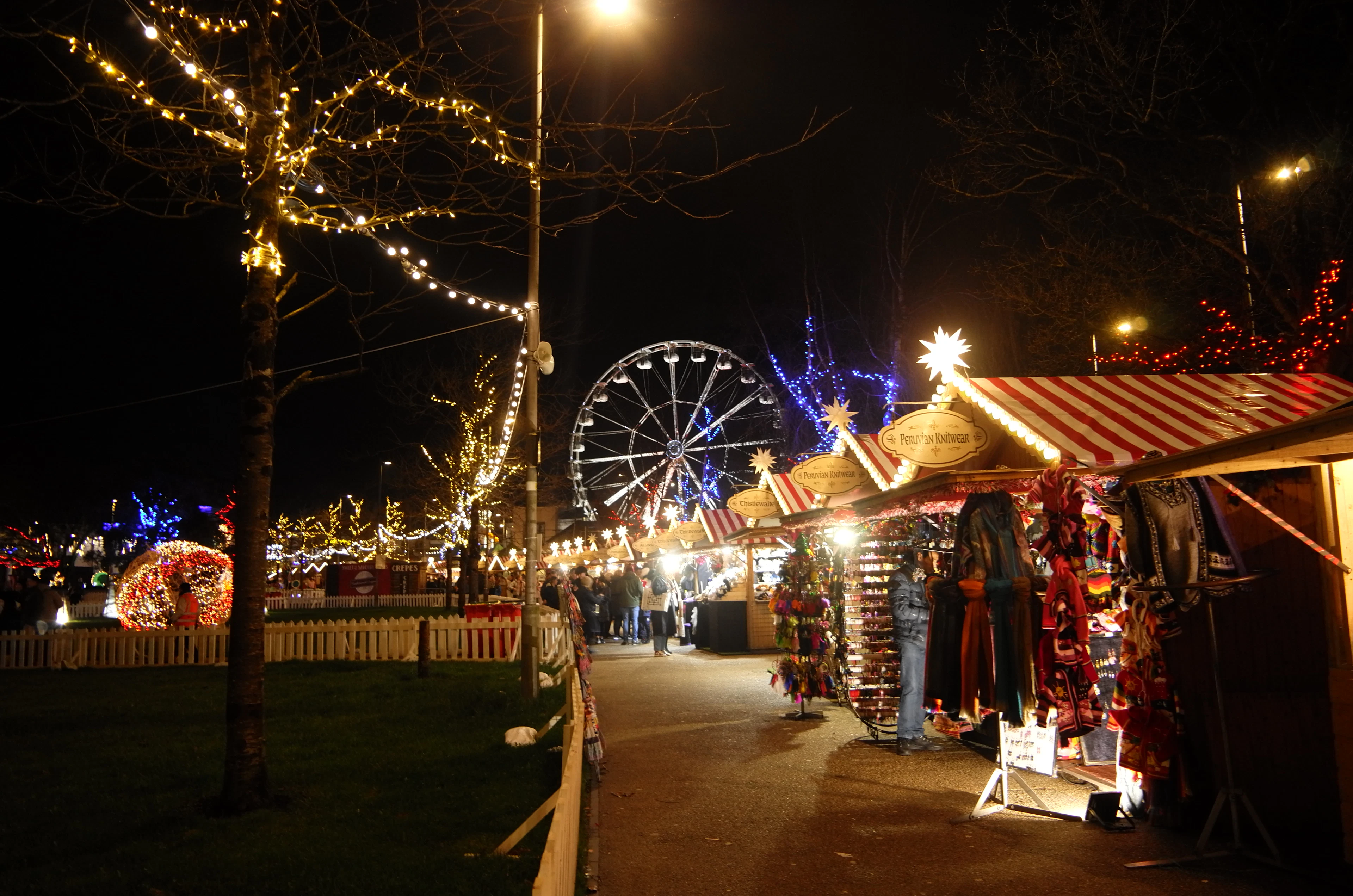 The Galway Christmas market in Eyre Square at night, red-and-white striped chalets strung with lights, fairy-lit trees along the path and the illuminated big wheel turning behind the stalls.